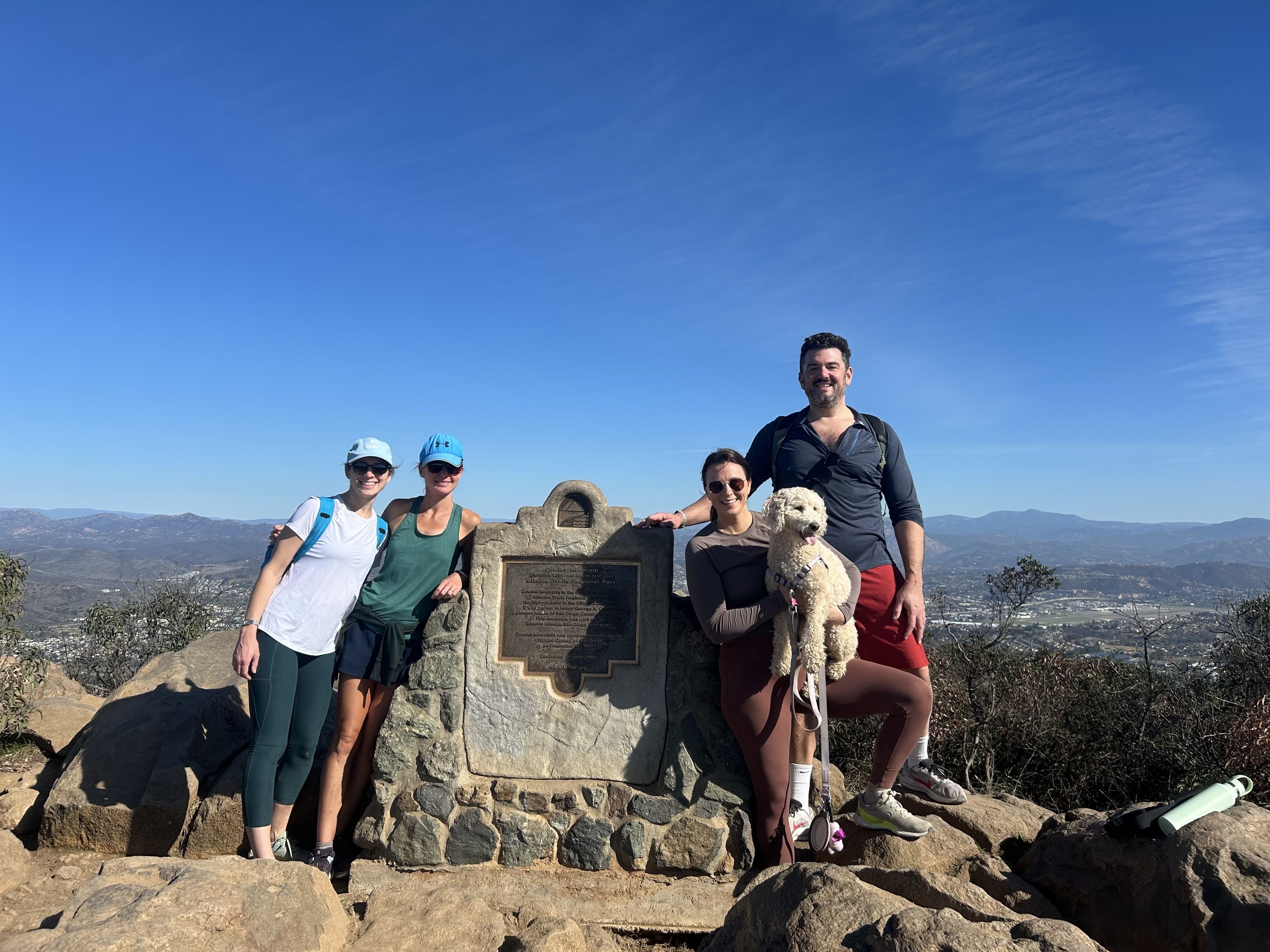 Four hikers and a fluffy white dog pose by a stone plaque on a rocky summit under a blue sky, with mountains and a town in the background. 
