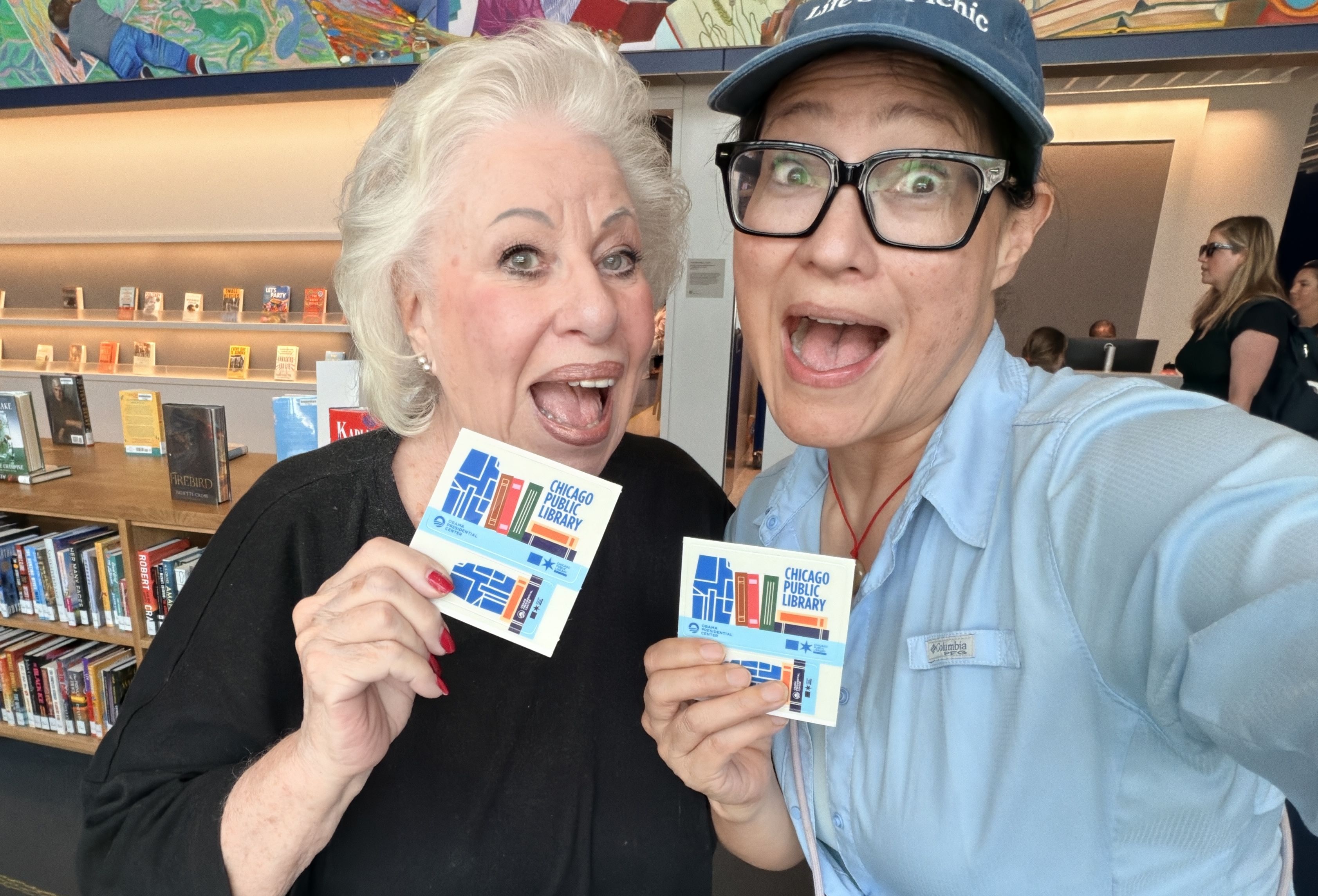 Two women pose for a selfie in a library, smiling; an older woman with white hair and a black top stands beside a younger woman in a blue cap and glasses; they hold Chicago Public Library cards.