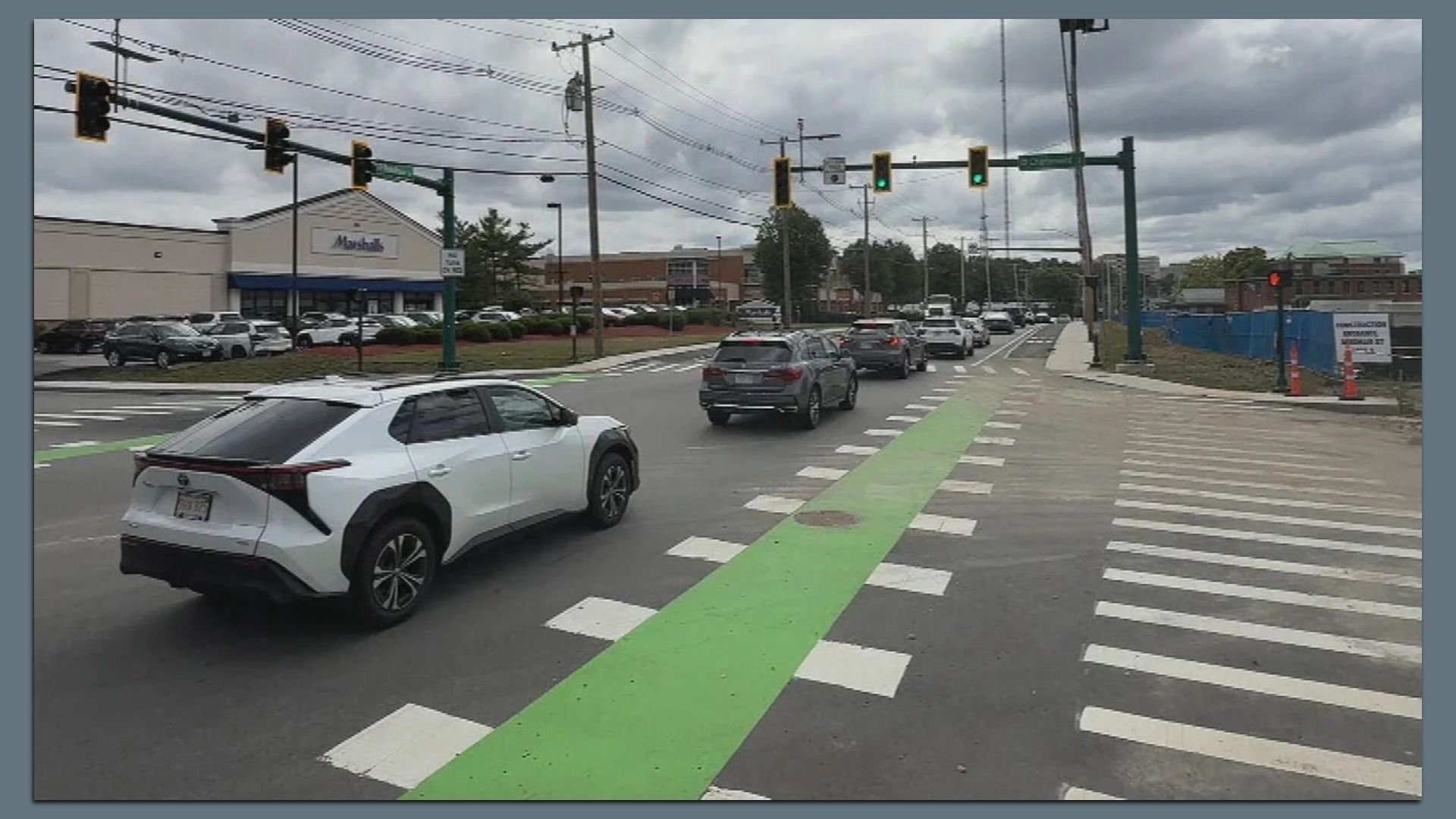 The intersection of Needham and Charlemont streets with traffic lights, multiple cars stopped or moving, a green bike lane, crosswalks, a Marshalls store on the left, and cloudy sky overhead.
