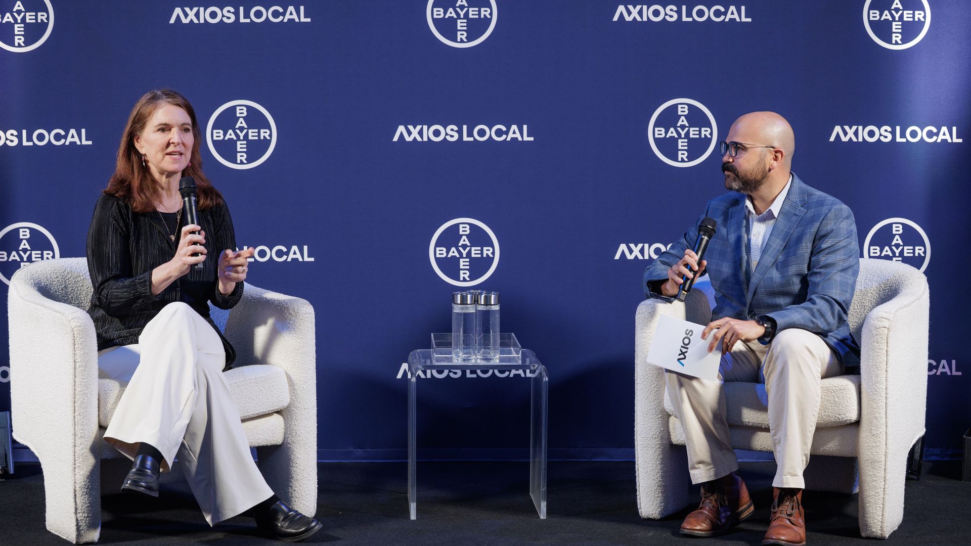 Two panelists sit on white chairs before a blue backdrop with the logos "AXIOS LOCAL" and Bayer logos; a woman on the left speaks into a handheld microphone, while a man on the right listens with a mic.