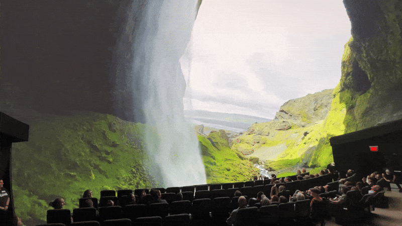 Indoor theater with an immersive floor-to-ceiling waterfall projection on a mossy green cliff backdrop, showing a scenic valley, while an audience sits and watches in dim lighting.
