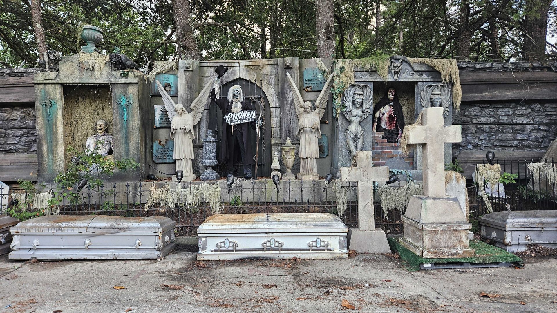 Haunted graveyard scene with weathered coffins, stone crosses, angel statues, and creepy figures including a skeleton holding a sign that says "Netherworld Haunted House" among tombstones and trees.