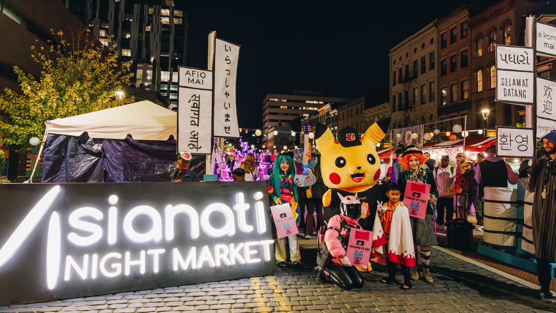 Night market at night featuring a yellow Pikachu mascot, people in colorful costumes, white vendor tents, string lights, and tall city buildings in the background.