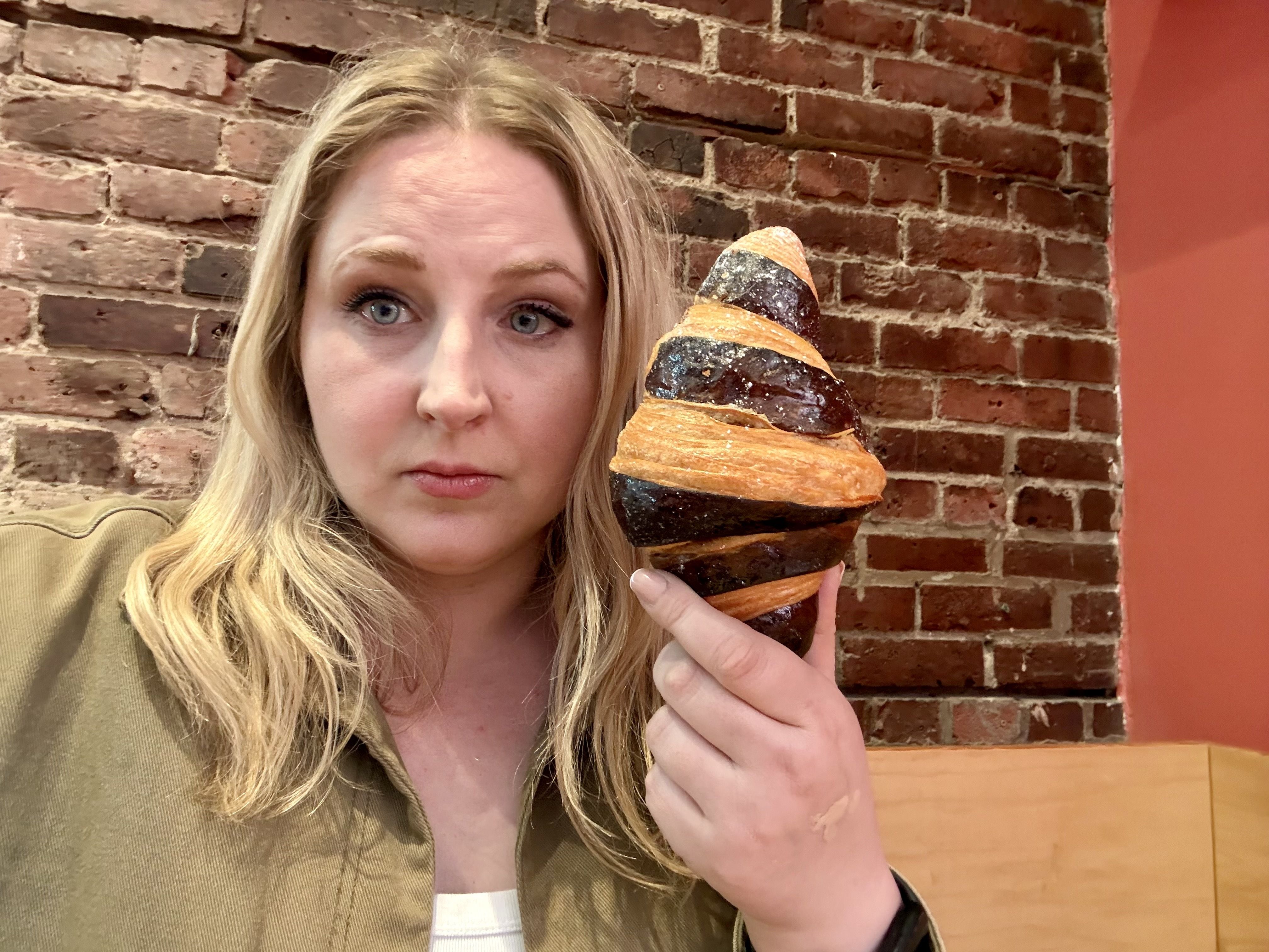 A woman with long blonde hair and blue eyes holds a large spiral pastry with orange and dark chocolate stripes, standing in front of a brick wall and looking at the camera.