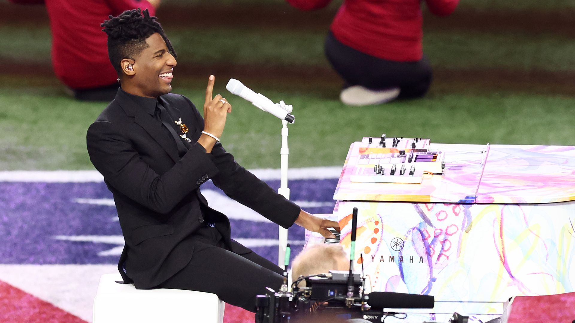 Jon Batiste holds up a finger and smiles as he plays a colorfully-painted piano.