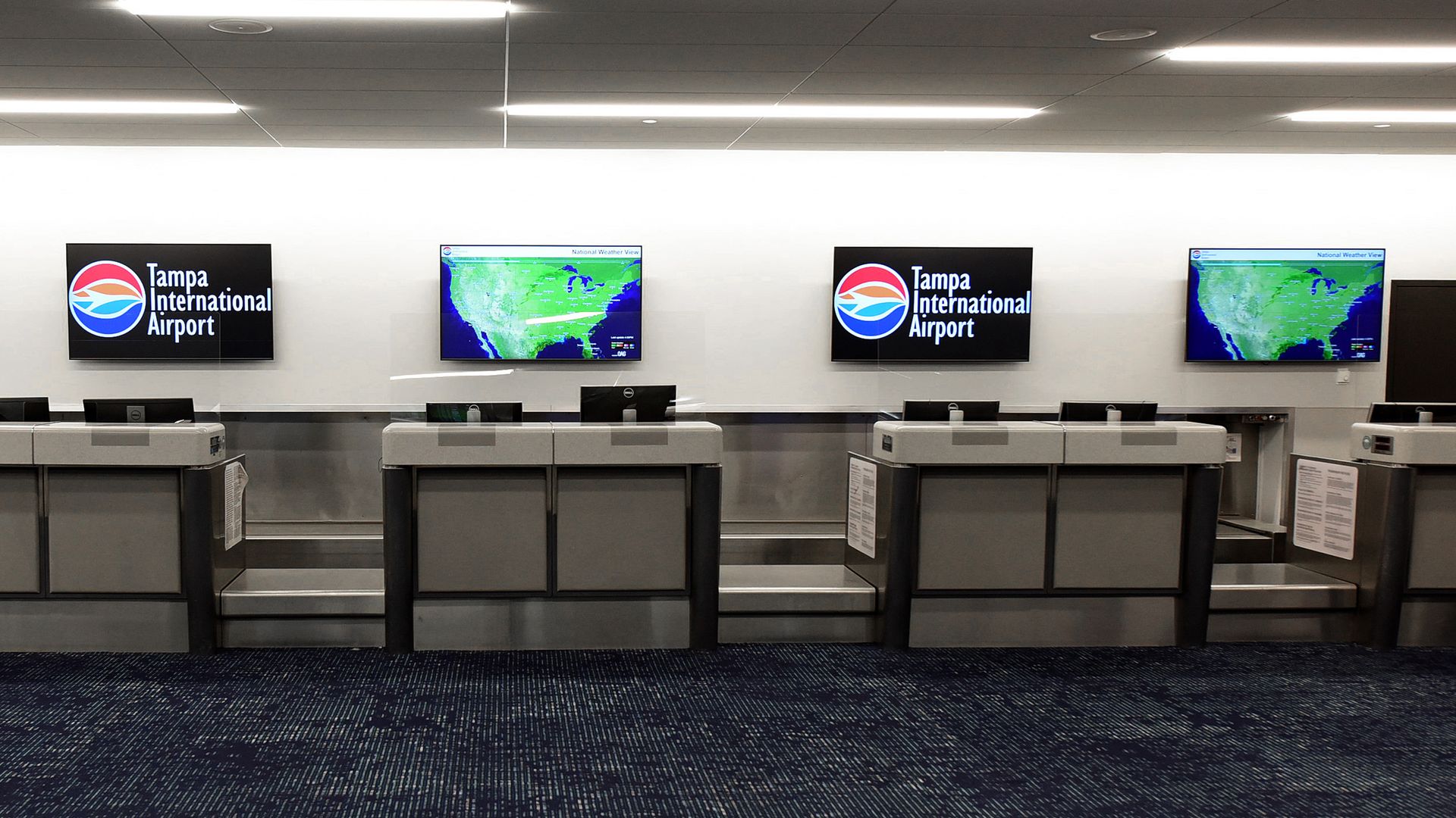 Wide view of a Tampa International Airport check-in area with four wall screens: two show the Tampa logo, two display weather maps; gray counters, barriers, and a dark blue speckled carpet.