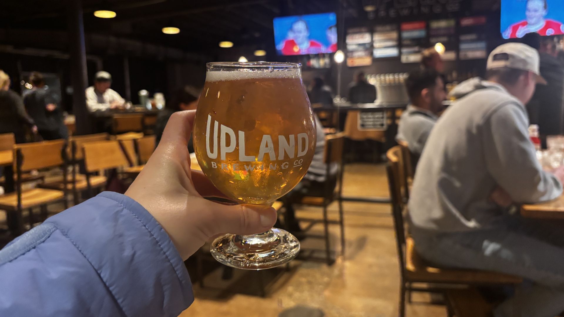Hand holding a glass of amber beer labeled UPLAND BREWING in a lively bar with wooden chairs and people watching sports on TVs.