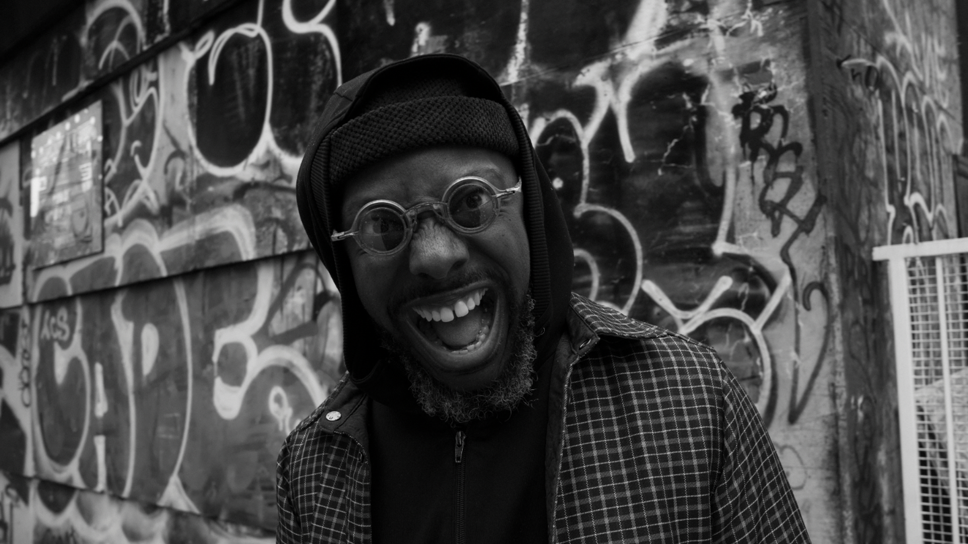 A man in a beanie and round glasses smiles in a funny and delightful way 