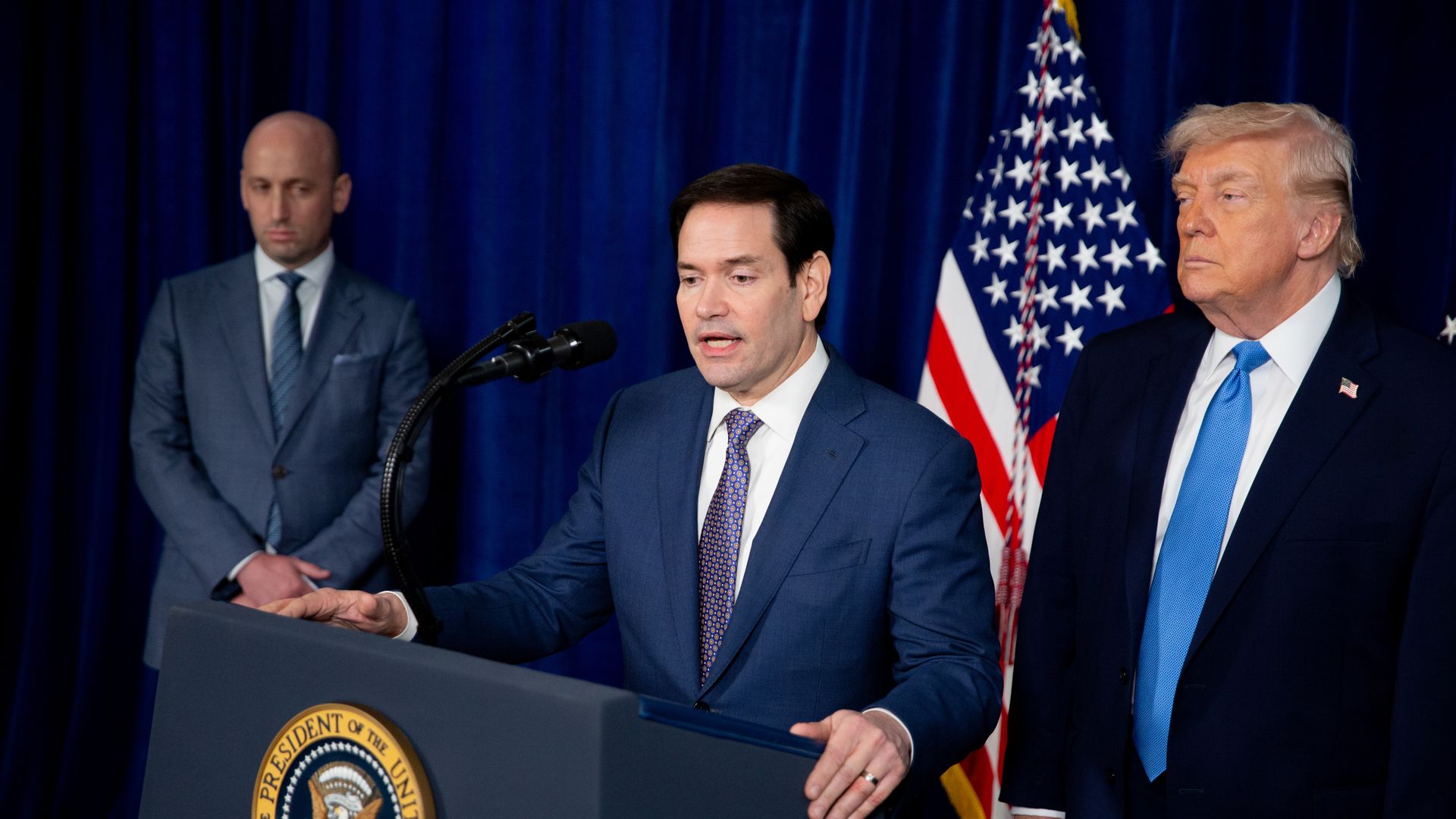Marco Rubio holds onto a podium while speaking into a microphone and is wearing a dark suit, a white collared shirt and a tie. President Trump and Stephen Miller flank him.