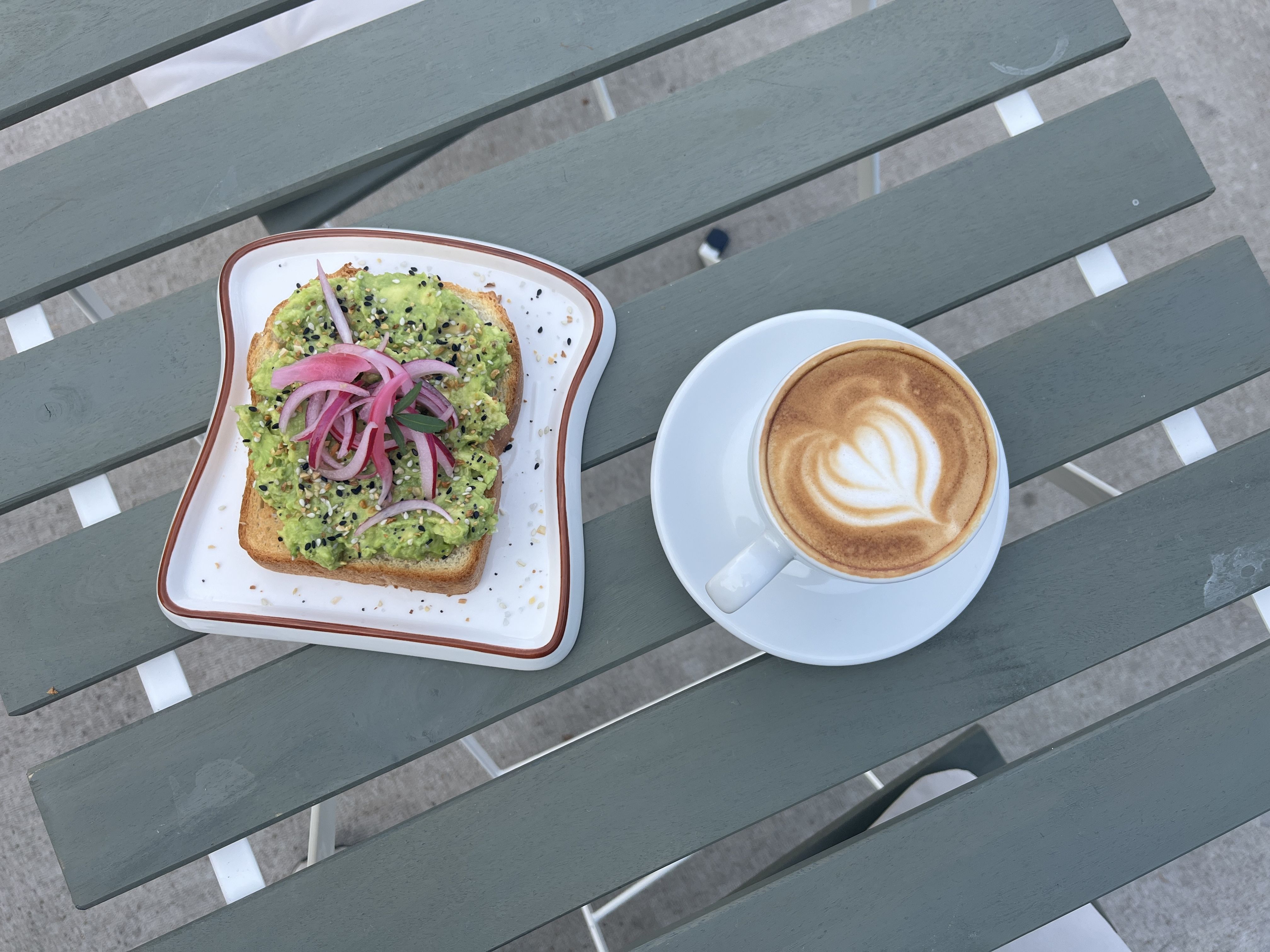 Avocado toast topped with sesame seeds and pickled red onions on a white plate with brown rim, next to a white cup of coffee with heart-shaped latte art, on a gray slatted table.