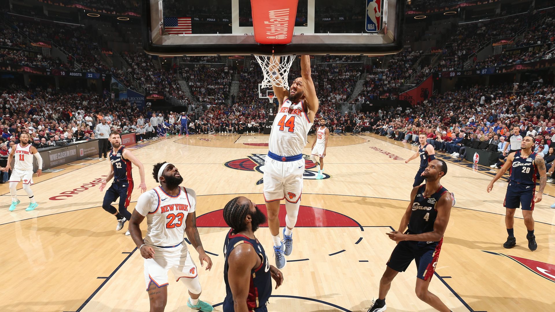 A Knicks player in a white jersey #44 dunks the ball, hanging on the rim as a packed arena watches; Cavaliers players and teammates stand around on the hardwood court.
