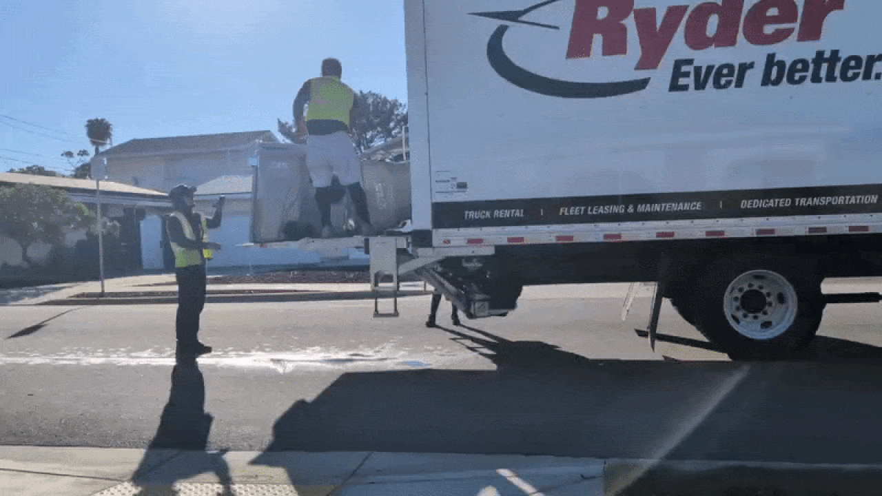 Two workers in neon vests load a Ryder white semi-trailer on a residential street: one on the trailer ramp inspecting, another on the ground signaling; bright sun, long shadows, and a clear blue sky.