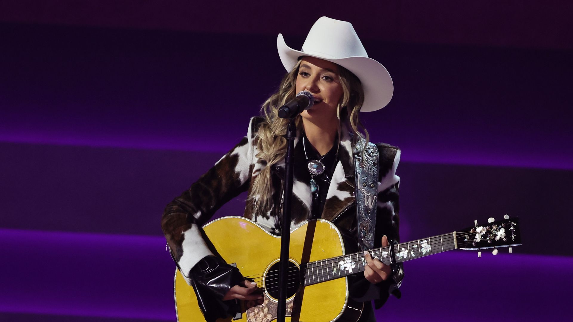 Lainey Wilson performs onstage in a cowboy hat and animal print jacket.