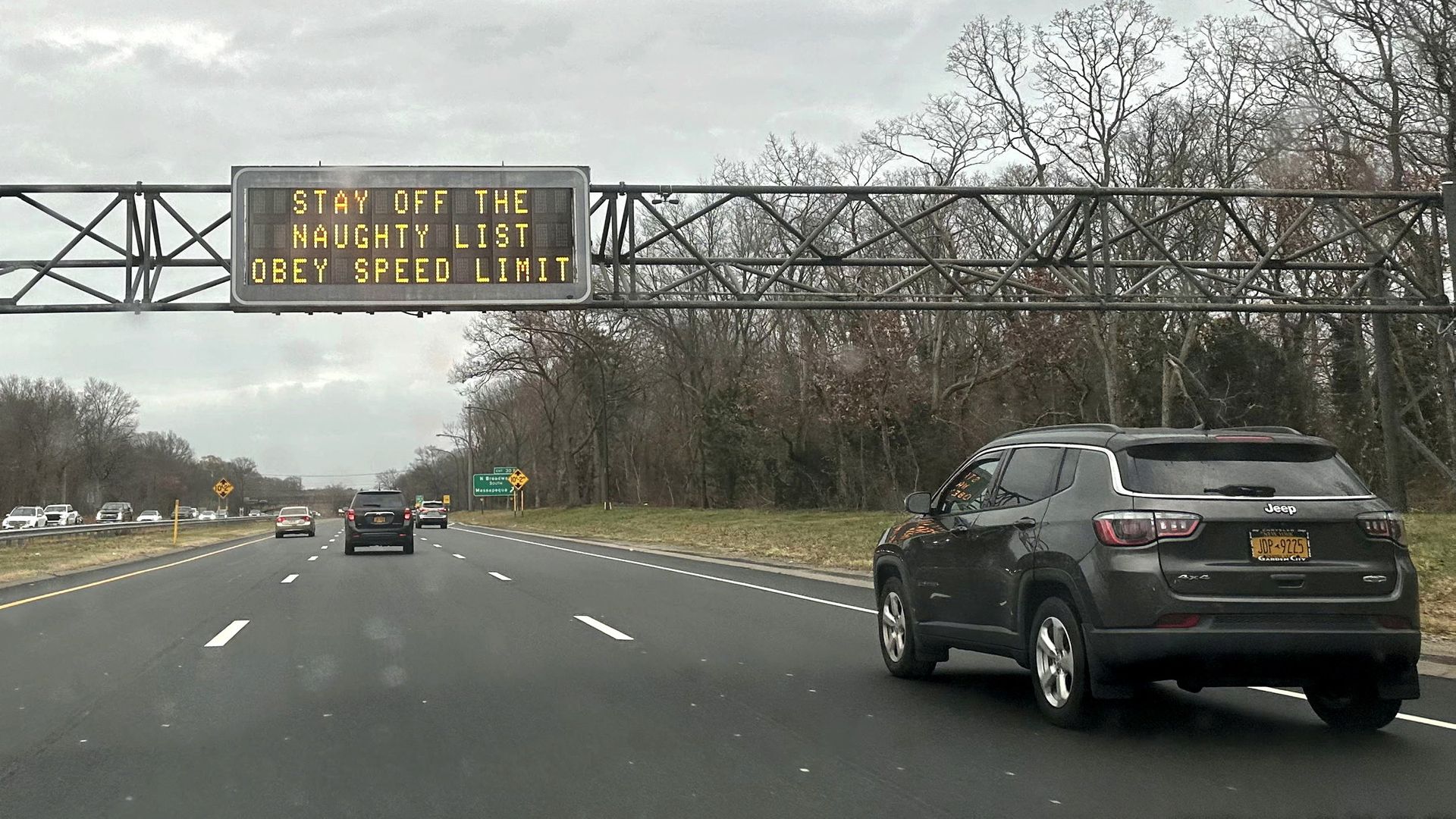 Highway scene with cars driving under an electronic sign that reads, "STAY OFF THE NAUGHTY LIST, OBEY SPEED LIMIT," with bare trees and cloudy sky in the background.