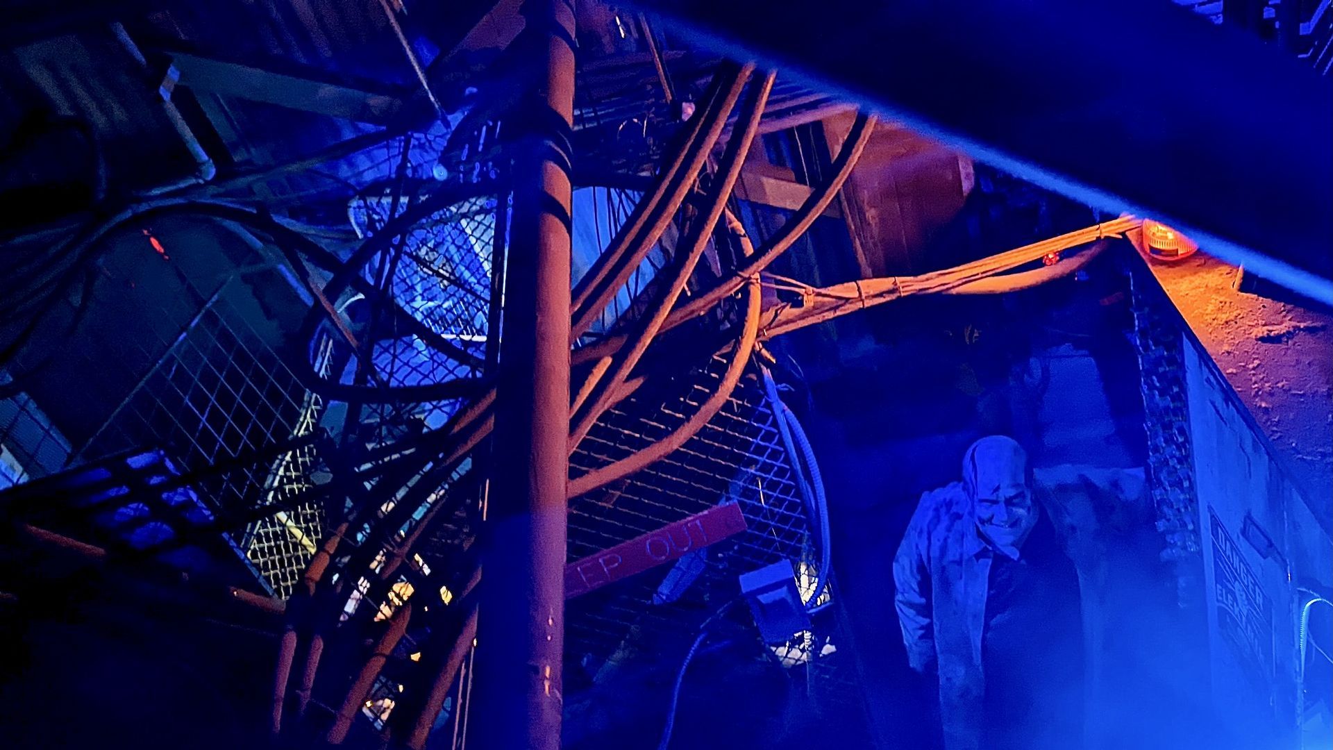 A man costumed as a scientist flashes an evil grin in a factory room with blue and orange lights and fog. 