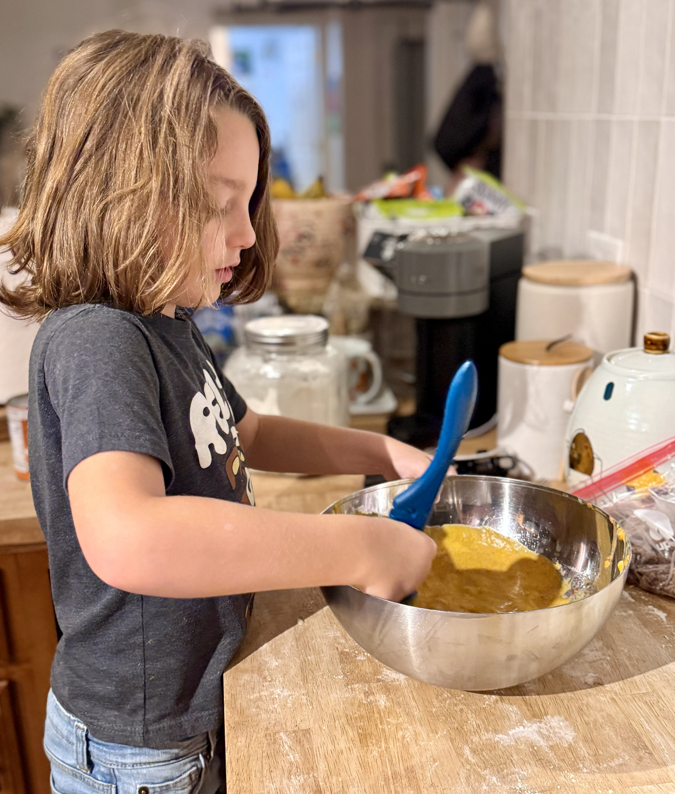 Child stirs orange batter in a silver mixing bowl on a wooden kitchen counter with baking supplies in the background.