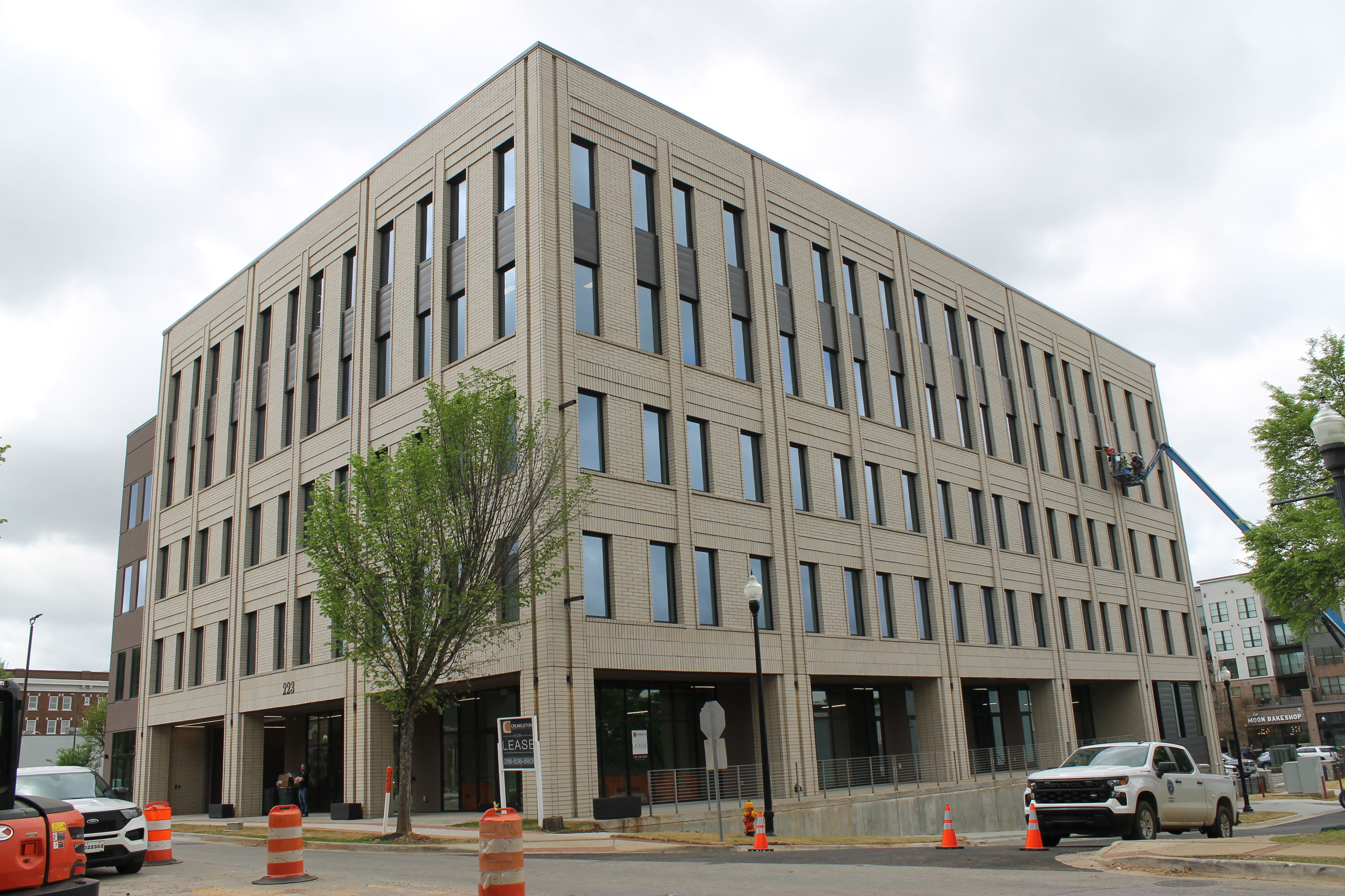 A beige brick office building with tall, narrow windows. A tree at the curb; orange barrels line the street, and a blue lift extends to the right near scaffolding; a white pickup is parked nearby.
