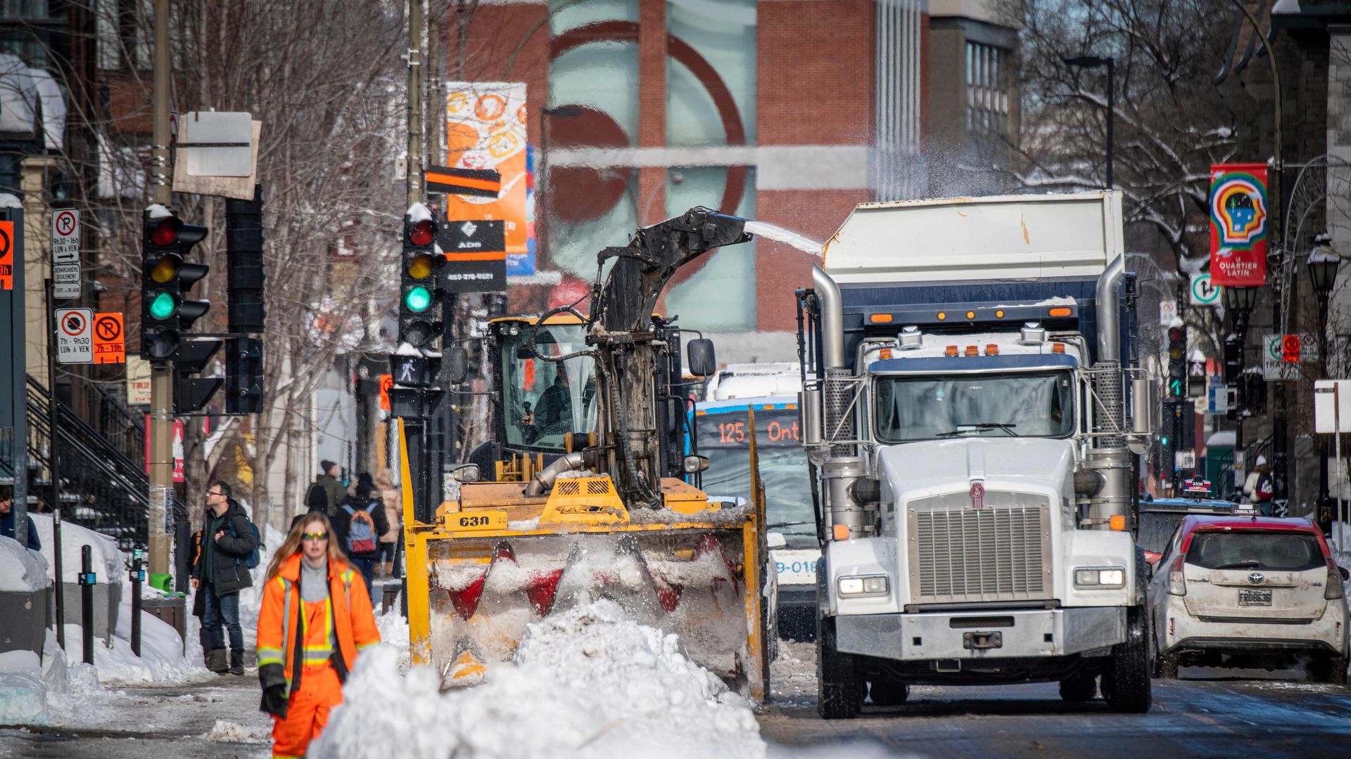 City street snow removal in progress with a worker in bright orange gear walking, a yellow snowplow pushing snow, and a white dump truck collecting it on a sunny winter day.