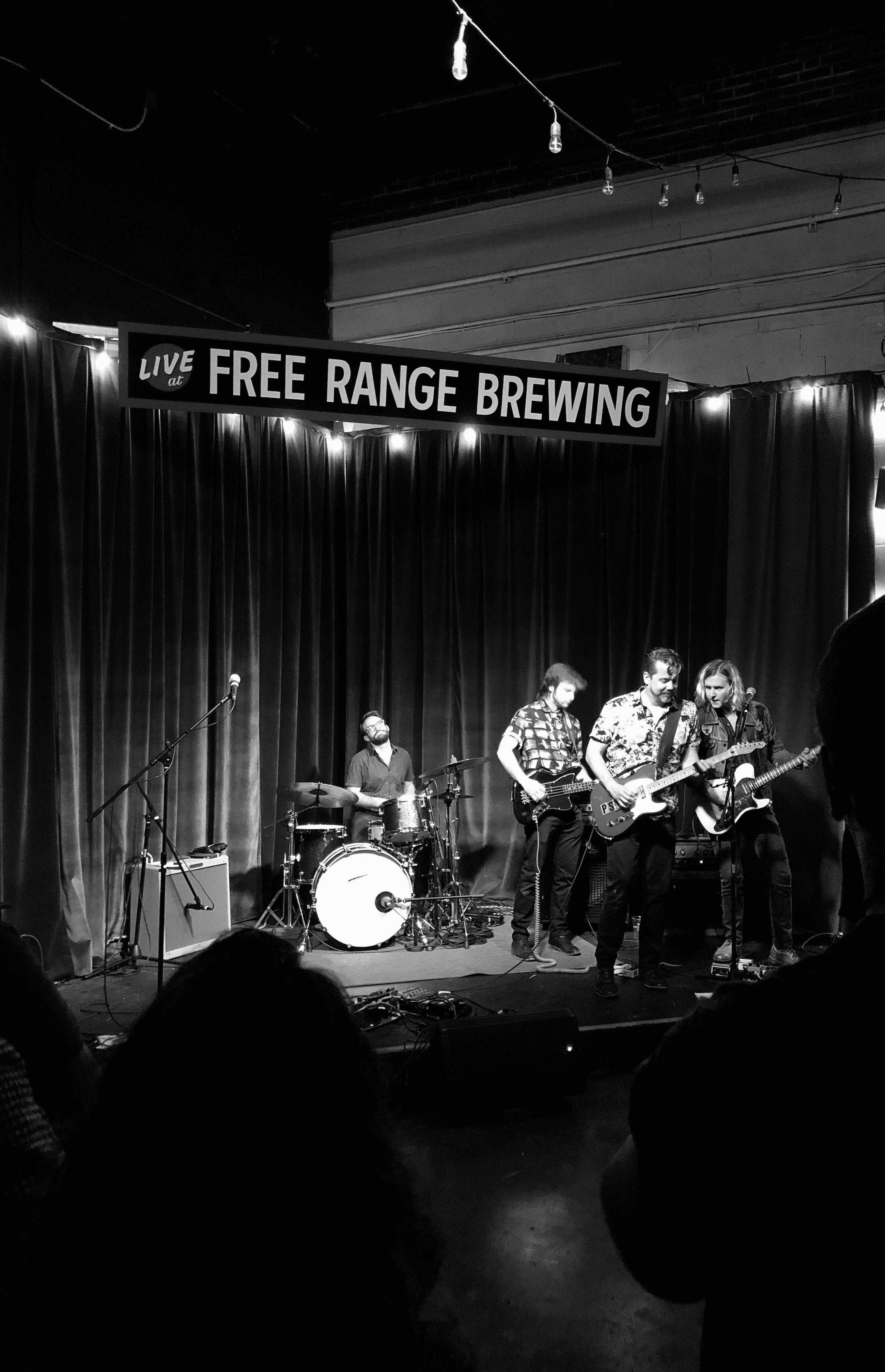 Black and white photo of a live band performance at Free Range Brewing with a drummer and three guitarists on stage against a curtain backdrop, illuminated by string lights overhead.