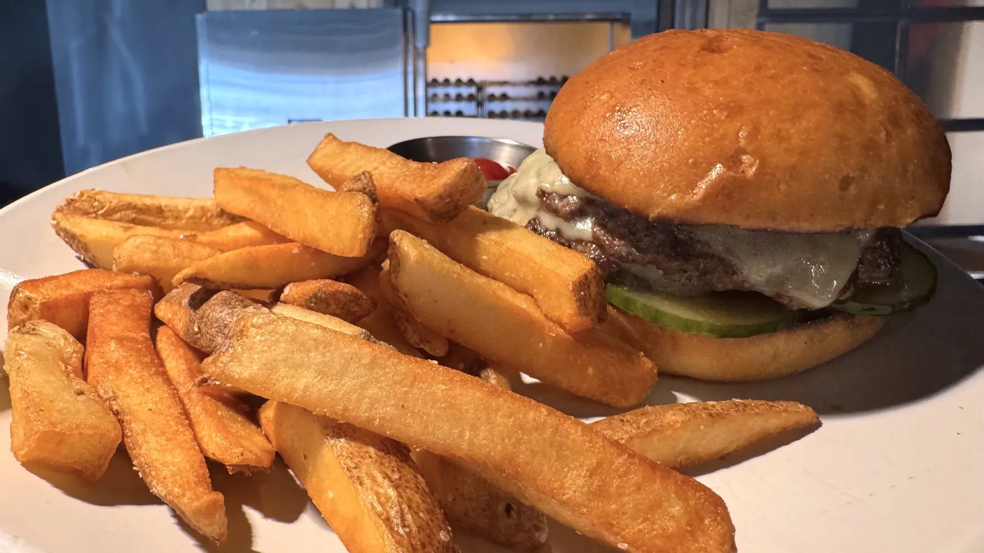 Plate with a cheeseburger and thick-cut fries: glossy golden bun, melted cheese oozing from the patty, pickle slice visible, in a kitchen setting.