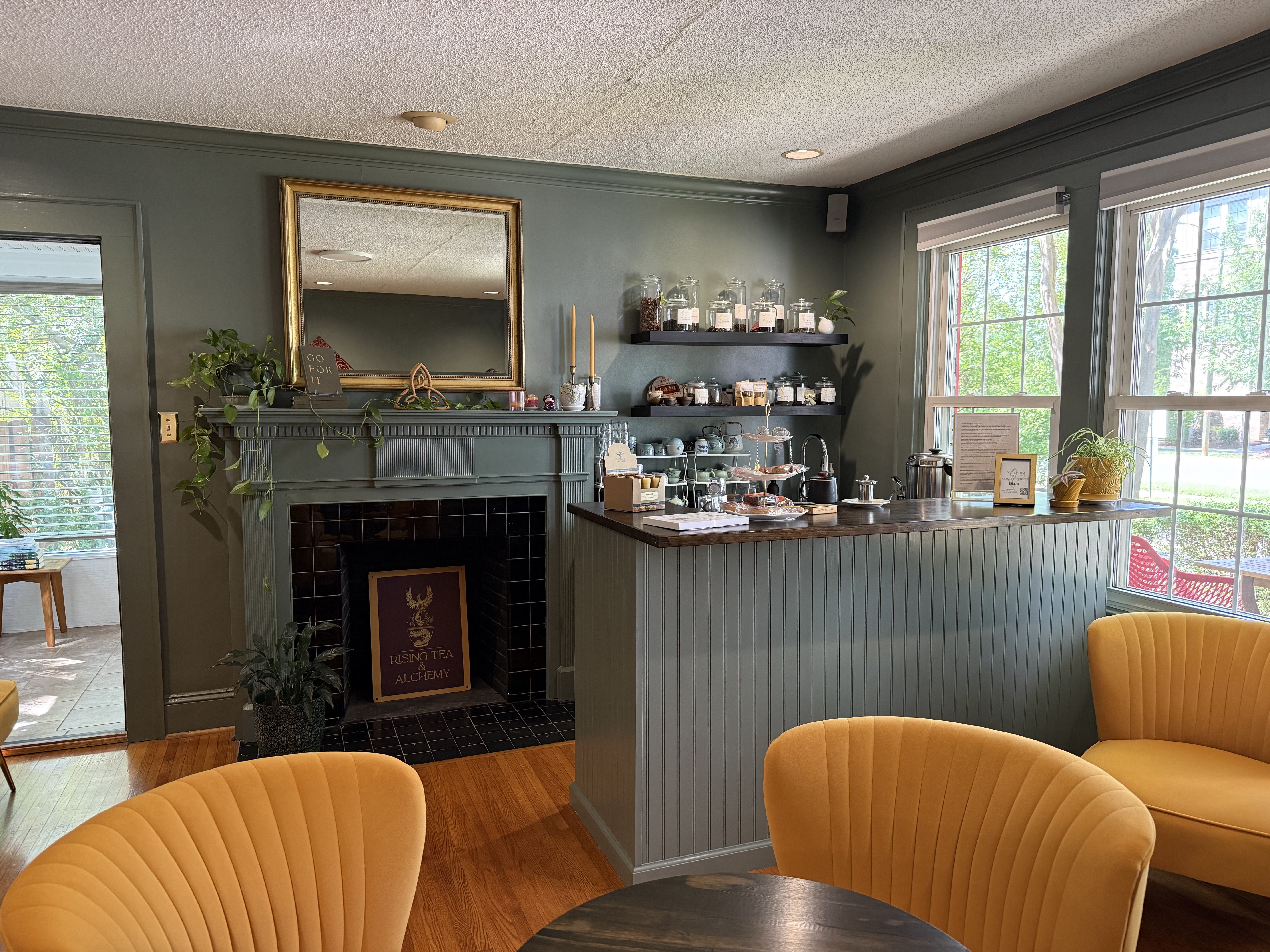 Cozy cafe interior with dark green walls, a fireplace mantel and gold-framed mirror, shelves of glass jars, potted plants, and bright yellow curved chairs around a wooden table by large windows.