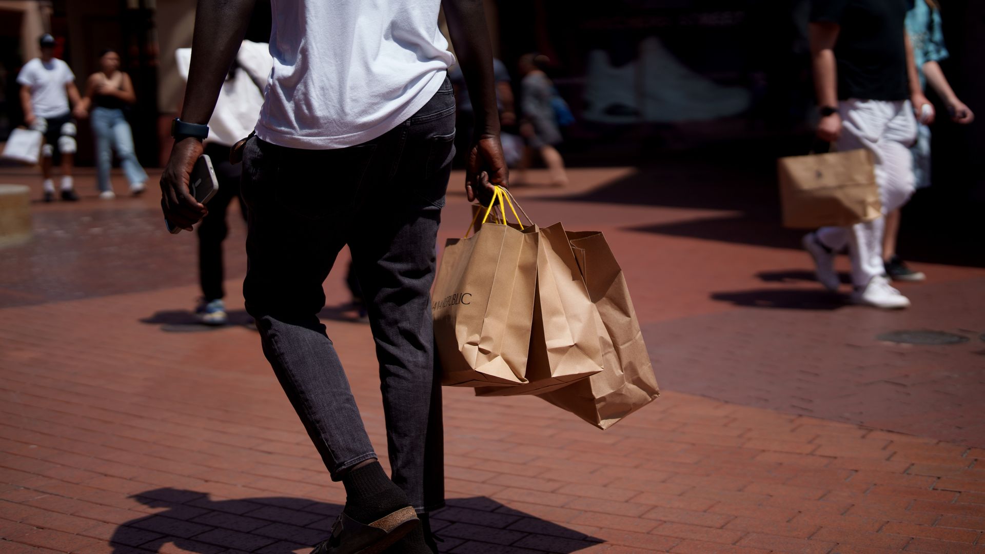 A person in black pants and a white shirt carrying brown paper shopping bags with yellow handles while walking on a sunlit brick pavement, other pedestrians blurred in background.