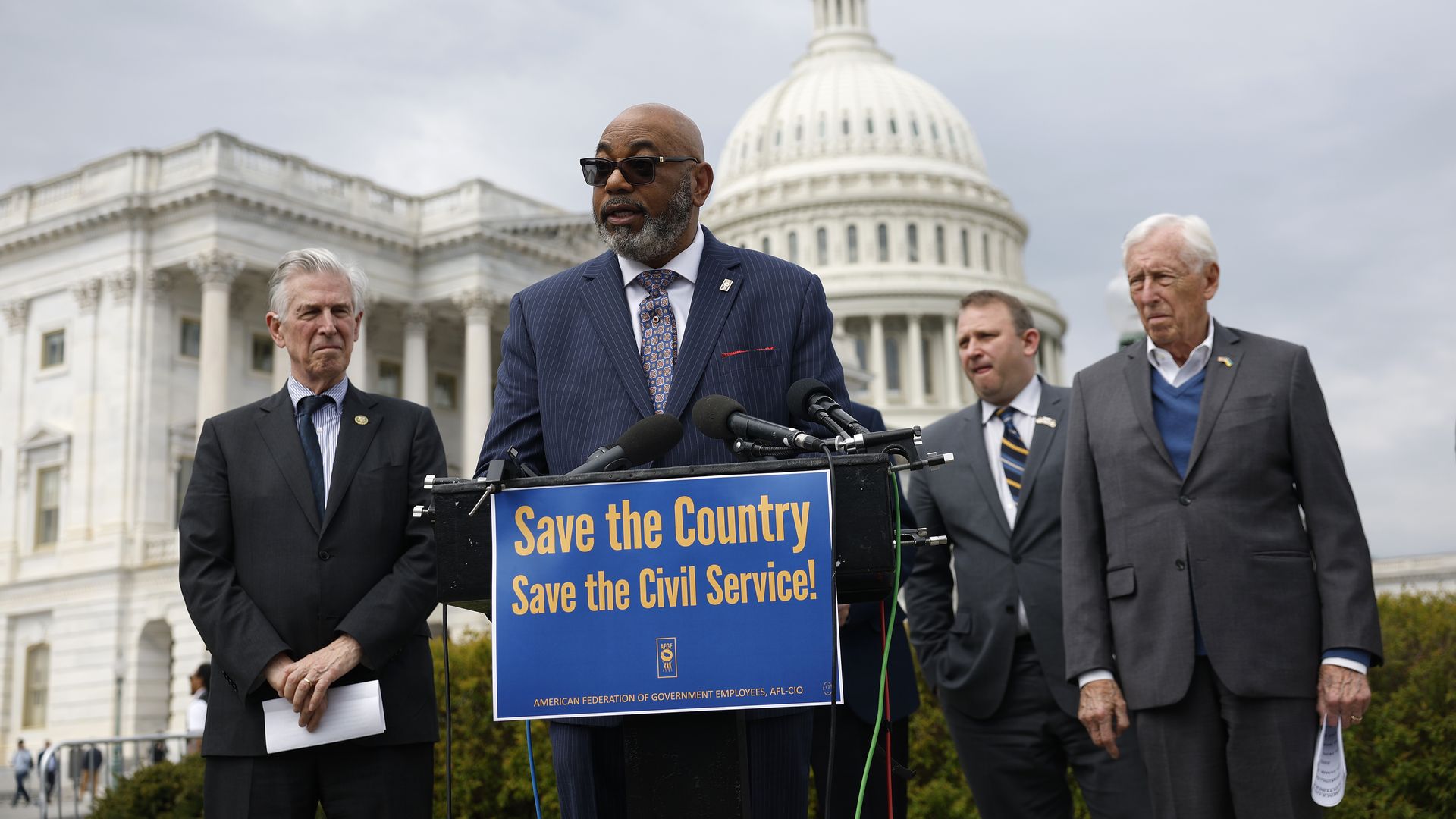 Everett Kelley, wearing a pinstriped blue suit, speaks at a podium with a blue "save the country, save the civil service!" poster on it while surrounded by House Democrats and standing in front of the U.S. Capitol building.
