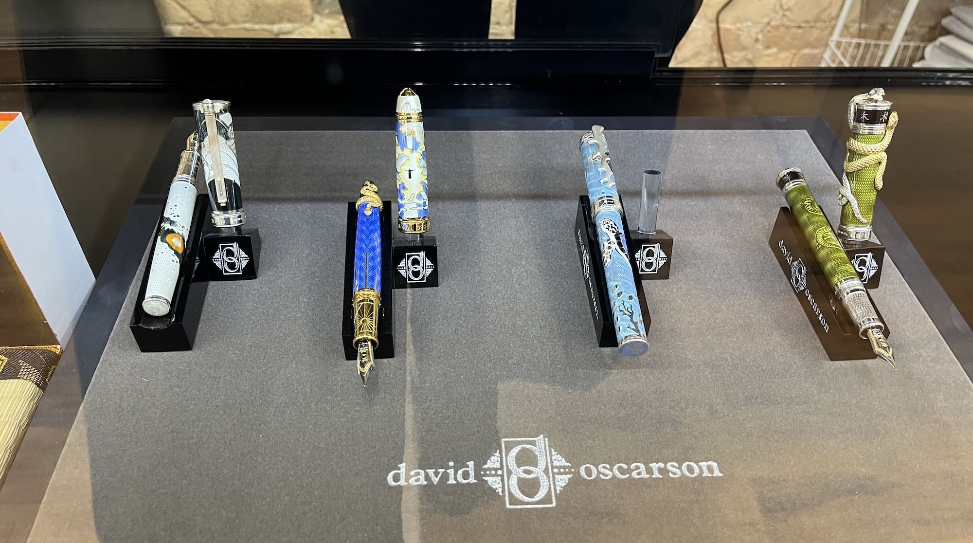 Display of four decorative fountain pens with intricate designs in light blue, dark blue, sky blue, and green, each on individual black stands with "david oscarson" branding.