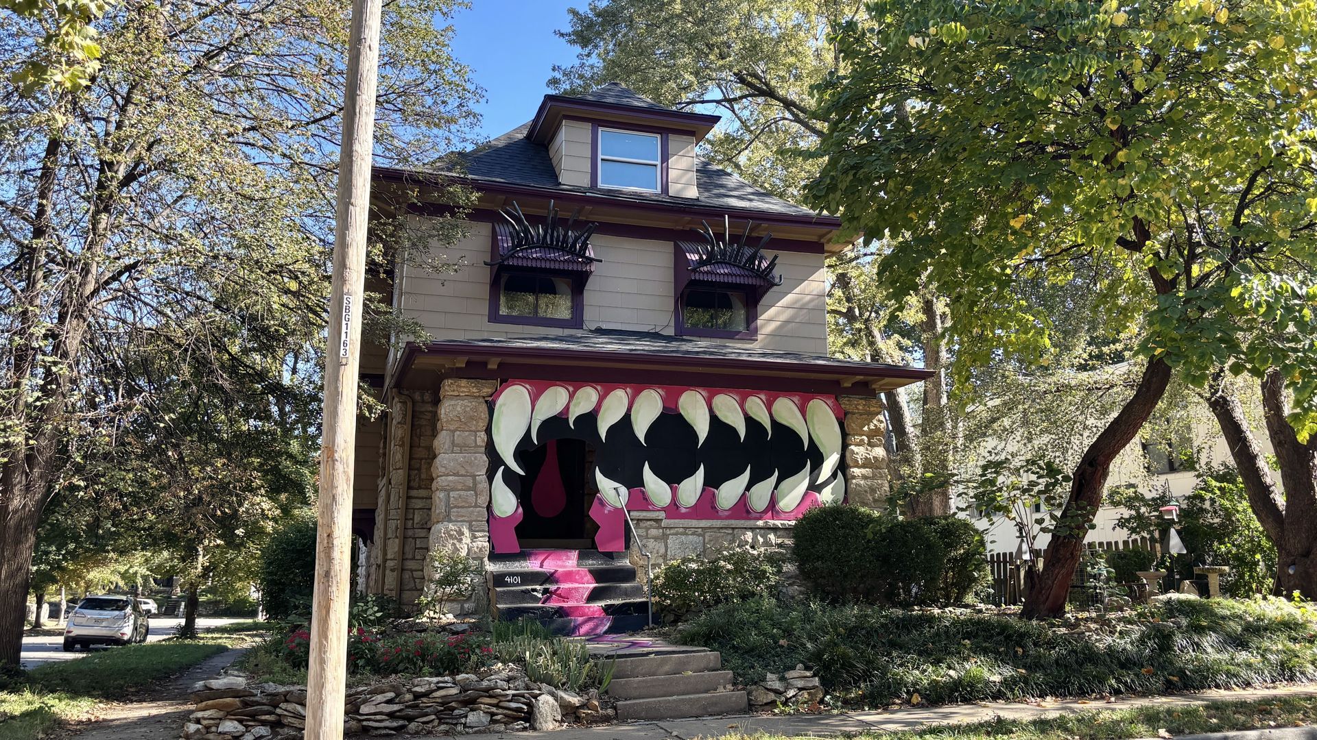 Two-story house with beige siding and purple trim, dormer window on top, and whimsical mural of a giant mouth with white teeth around the front doorway. Stone base, lush trees surround.