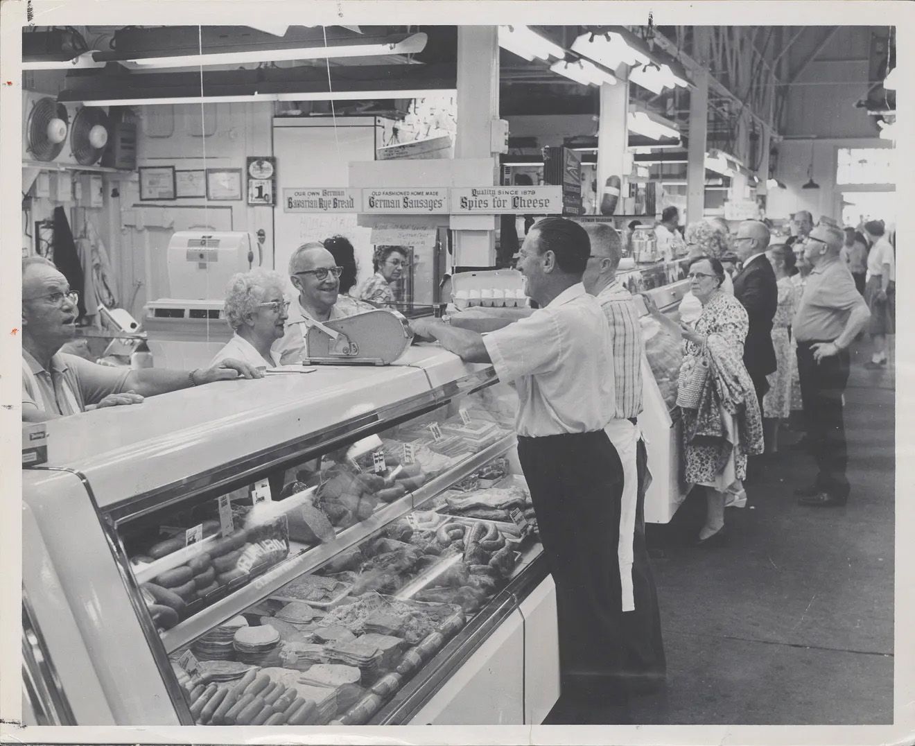 Black-and-white photo of a busy meat counter with a glass display at Findlay Market in 1963; customers line up as a worker weighs meat. Signs read "German Sausages" and "Spices for Cheese".