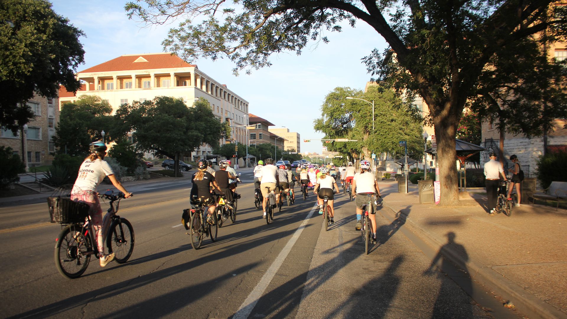 Crowd of cyclists rides down a sunlit city street, lined with green trees and red-tiled roofs on midrise buildings. Foreground riders pedal away as long shadows stretch across the pavement.
