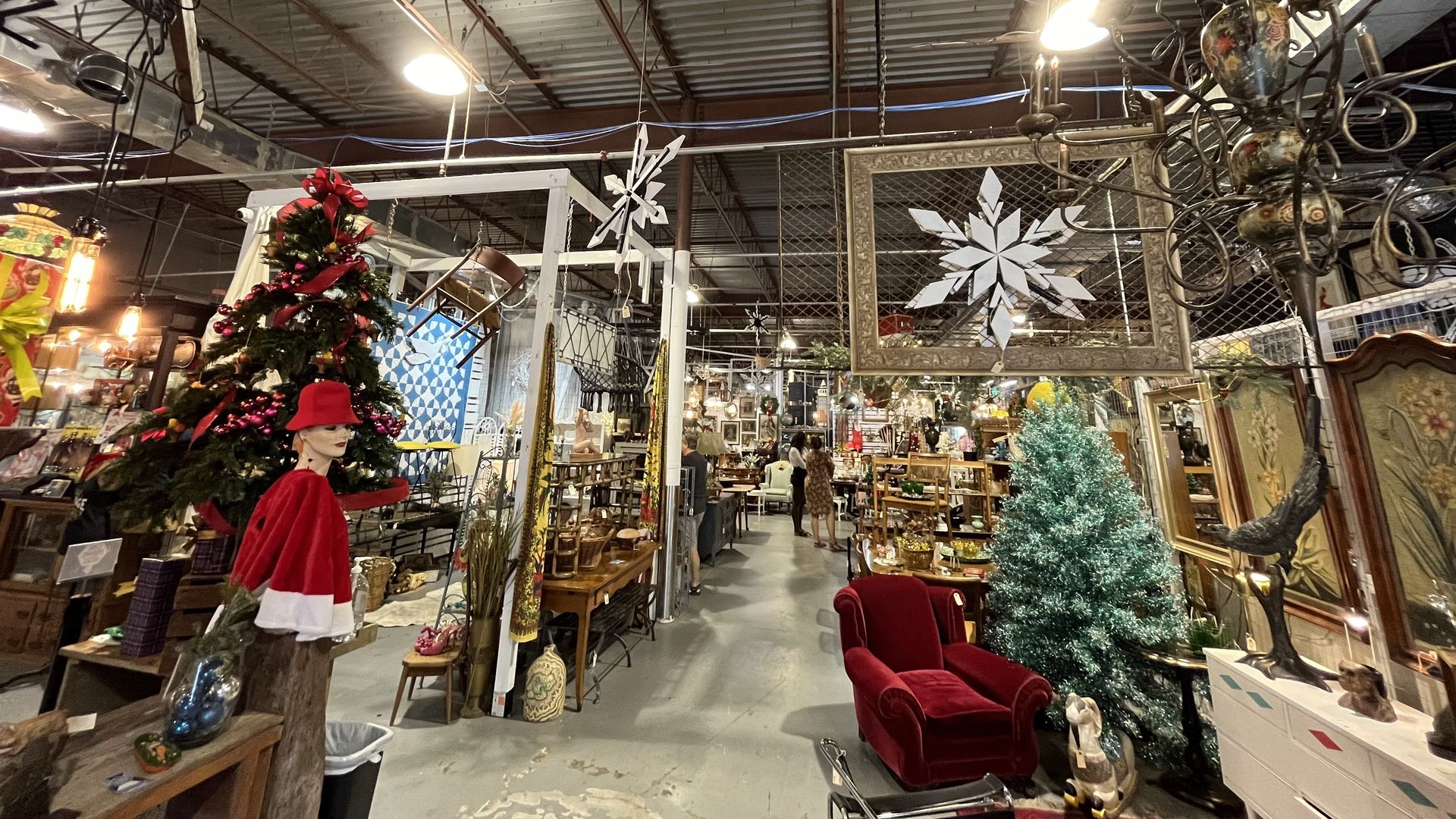 Antique store with Christmas decorations including a green tree with pink ornaments, a teal tinsel tree, large white snowflakes, and a red velvet armchair amid vintage furniture and décor.