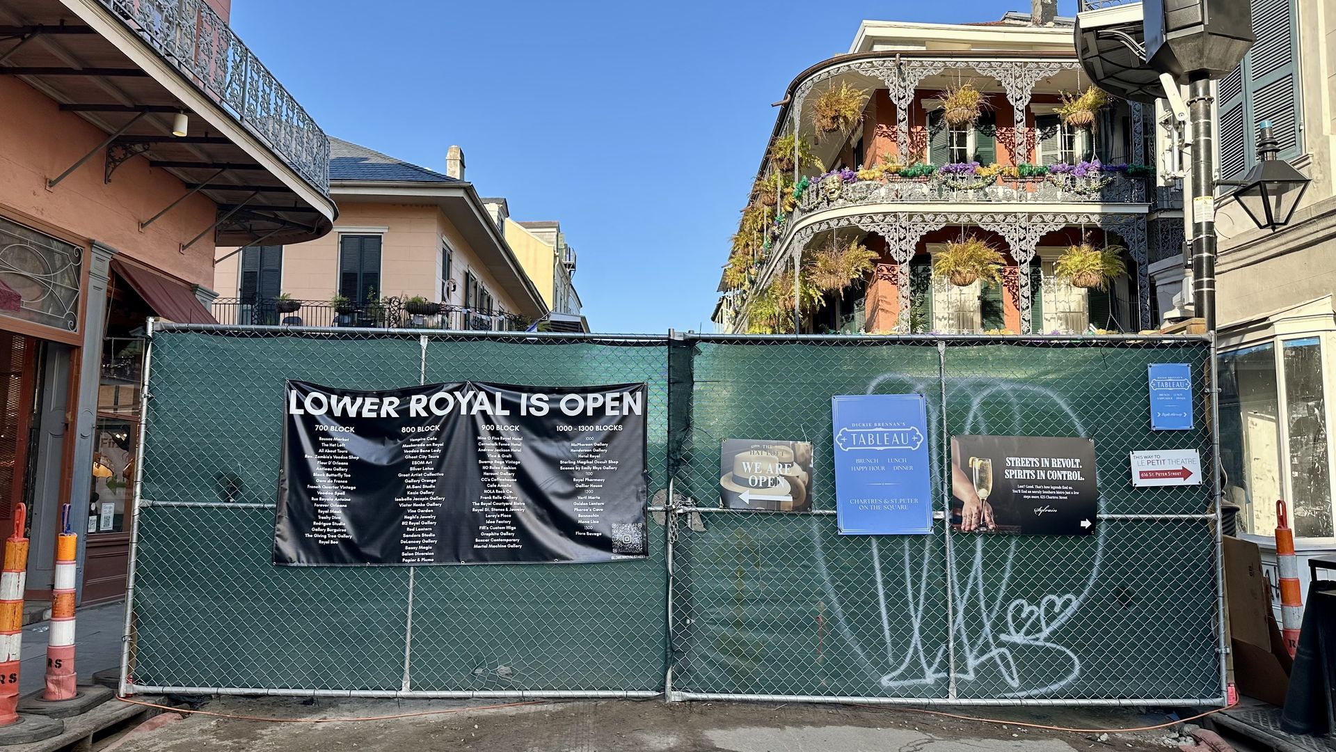 Green construction fence with signs reading "Lower Royal is Open," restaurant listings, and directions, set against ornate balconies with hanging plants under a clear blue sky.