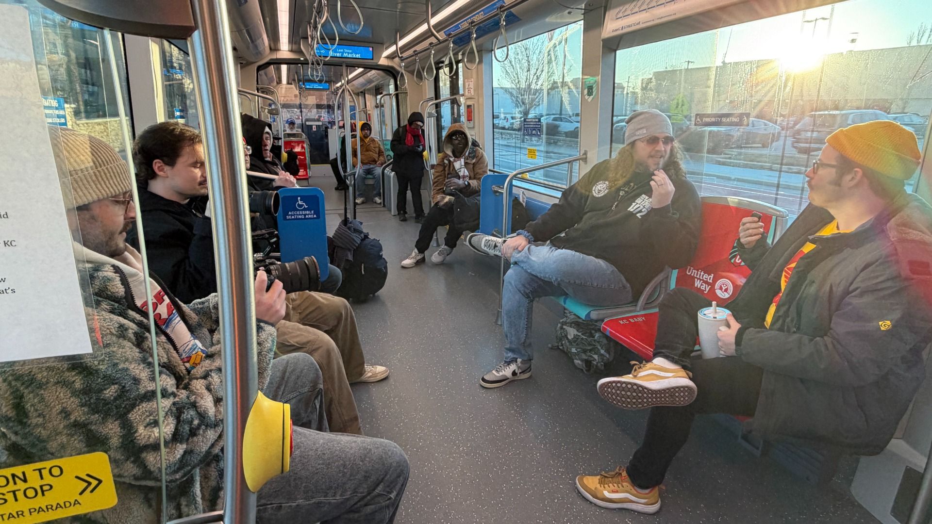 Inside a public transit vehicle, several passengers dressed in winter clothes sit and stand. Sunlight shines through large windows on the right side. Some passengers hold cameras or drinks.