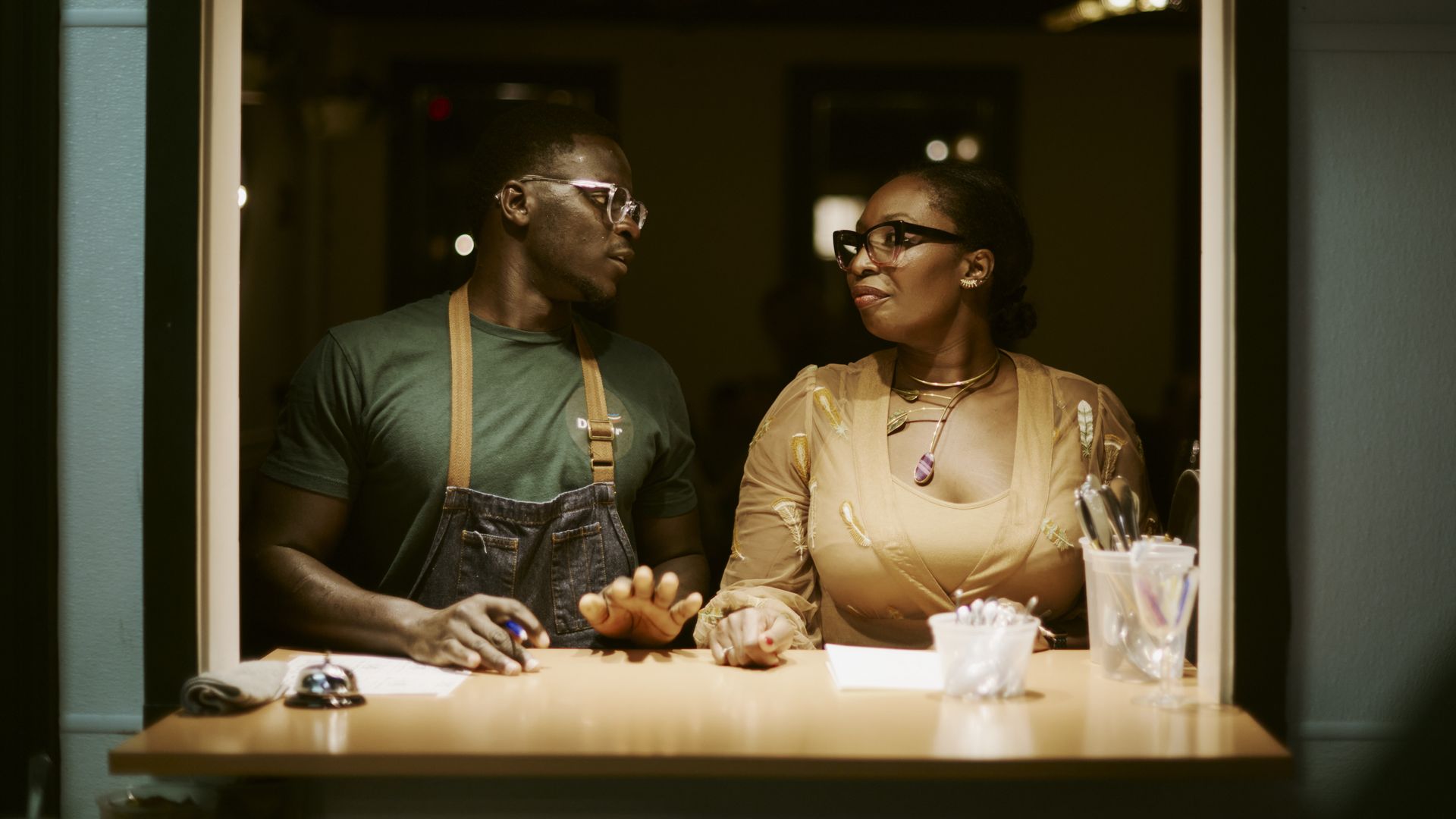 Serigne Mbaye and Effie Richardson pose for a photo at a service window inside Dakar NOLA. The moody lighting highlights the pair looking at each other as they stand in the window opening.