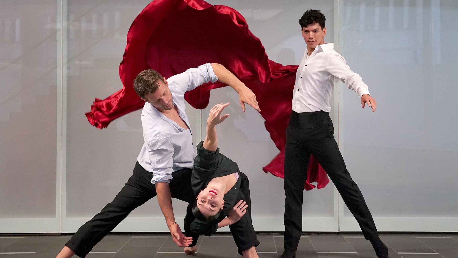 two men and a woman dance ballet while a large red silk cloth drapes behind them
