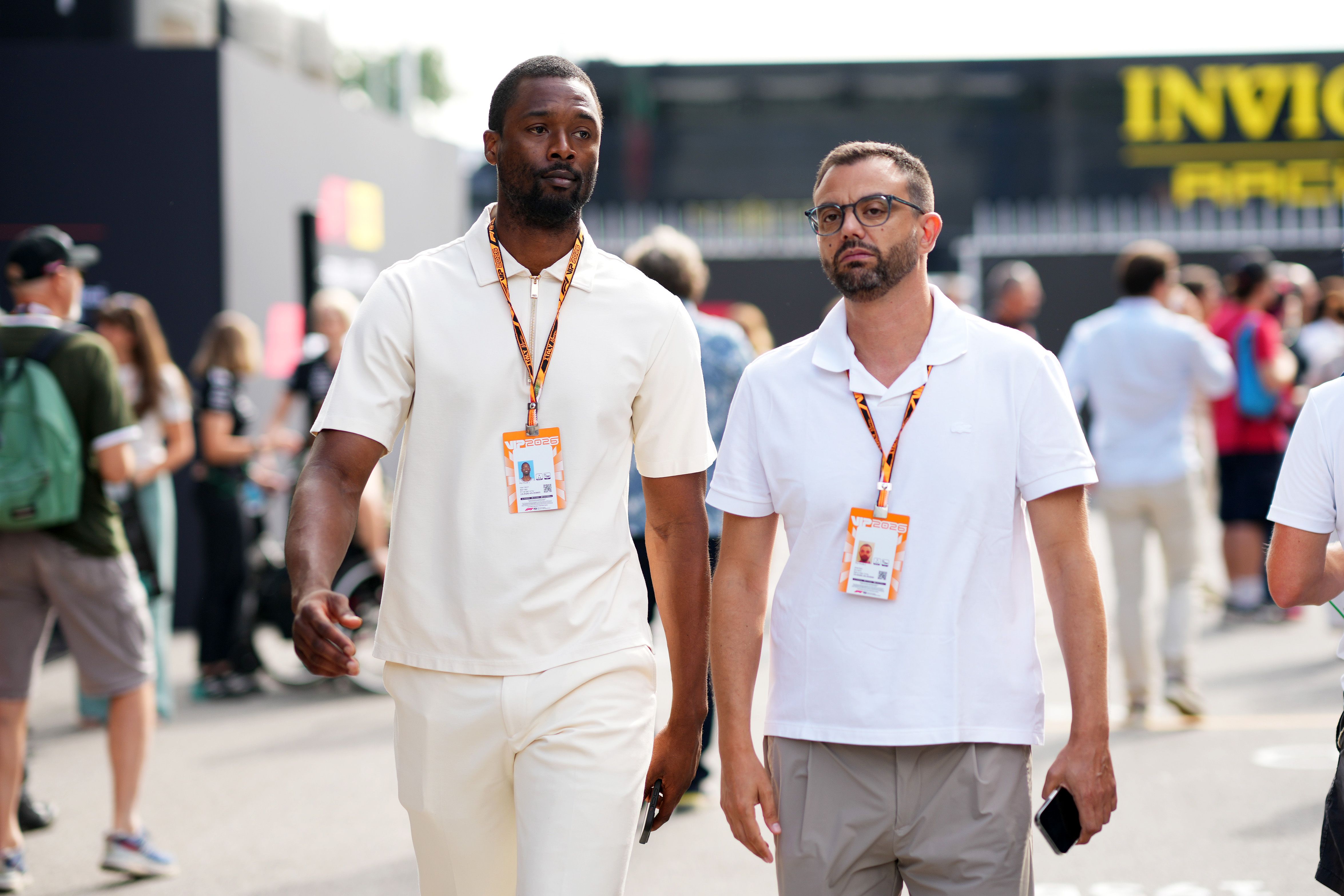 Two men with VIP badges walk at a crowded event. Left man wears cream polo and matching pants; right man wears white polo and gray shorts with glasses. Blurred attendees and signs in back.