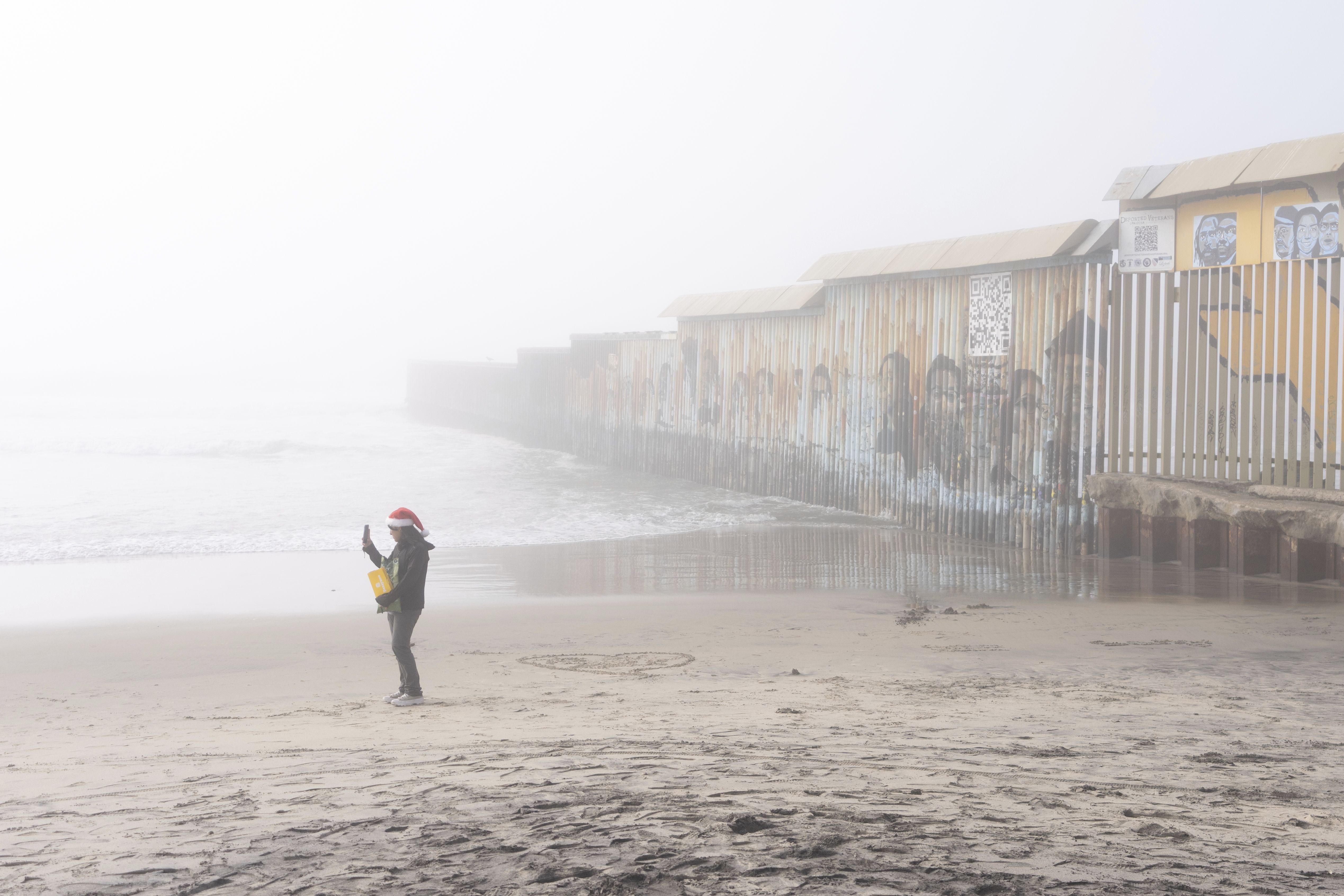 A woman wears a Santa hat as she takes a selfie next to the border wall separating Mexico and the United States in Tijuana.