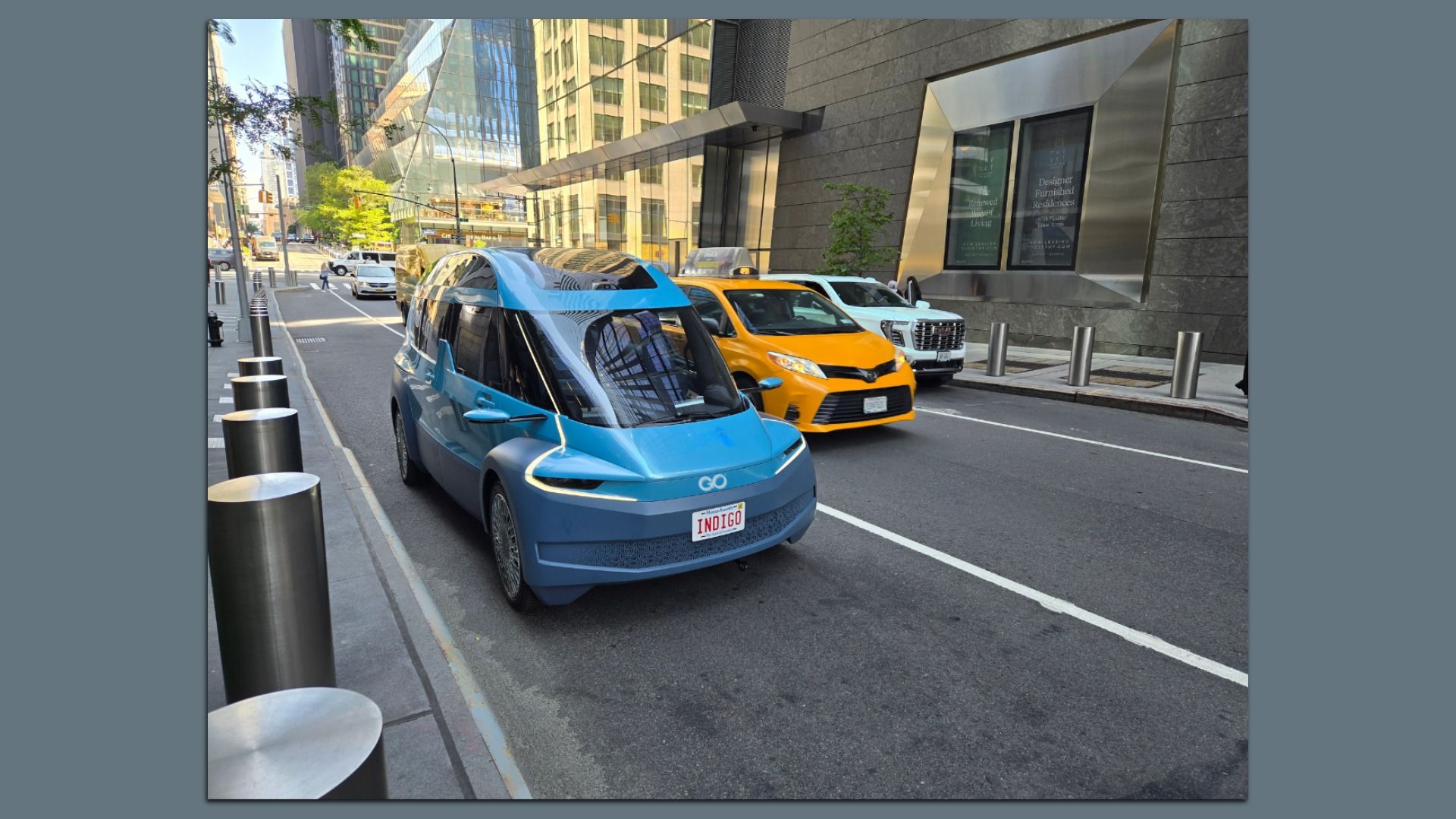 A blue Indigo Technologies electric van, designed for ride-hailing and delivery services, on a city street beside a yellow New York City taxi and a white pickup, with glass skyscrapers, a metal-framed storefront window, and metal bollards lining the curb.