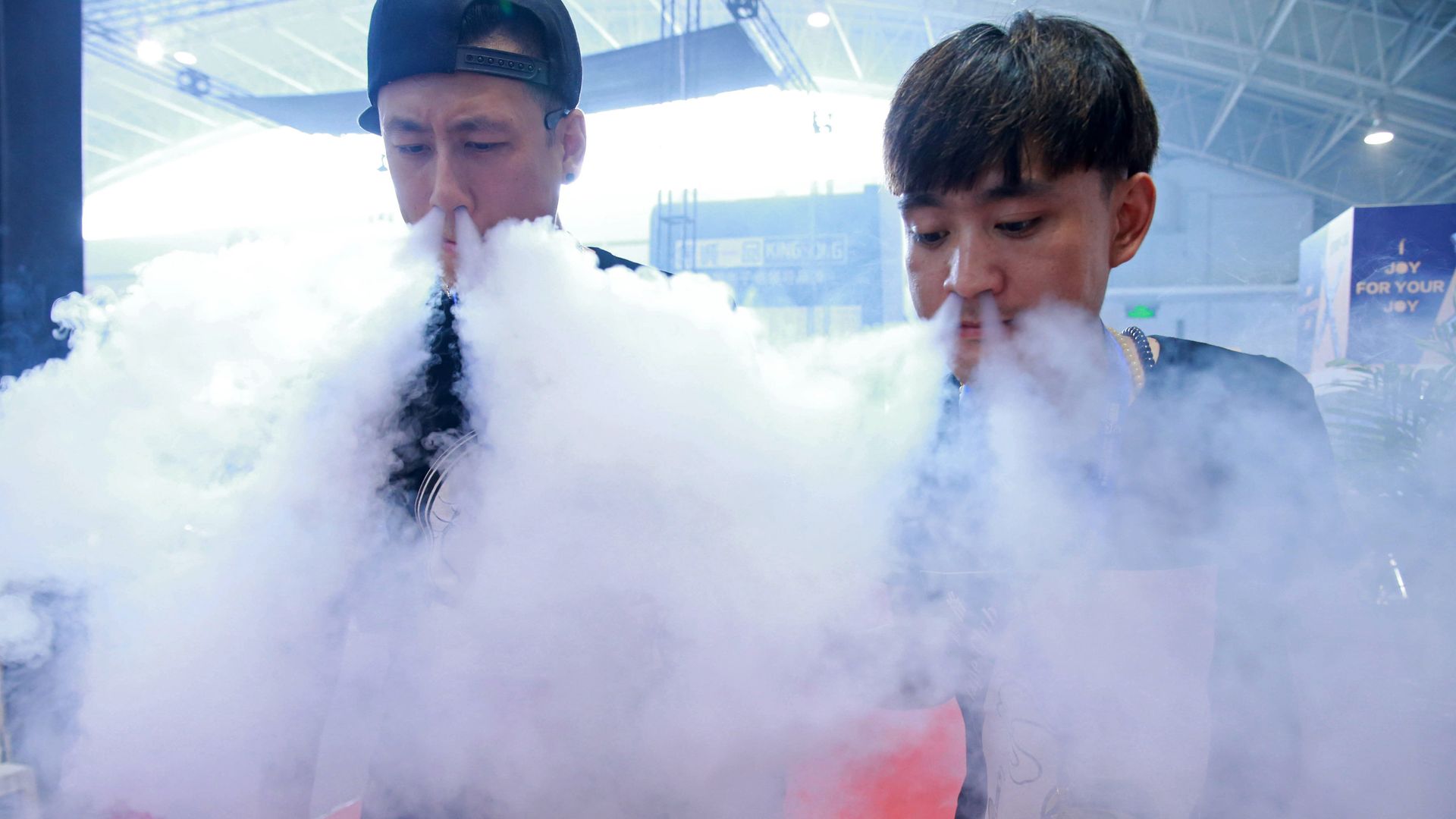 Two people at a Chinese expo showing how to vape, with lots of smoke coming out of their noses