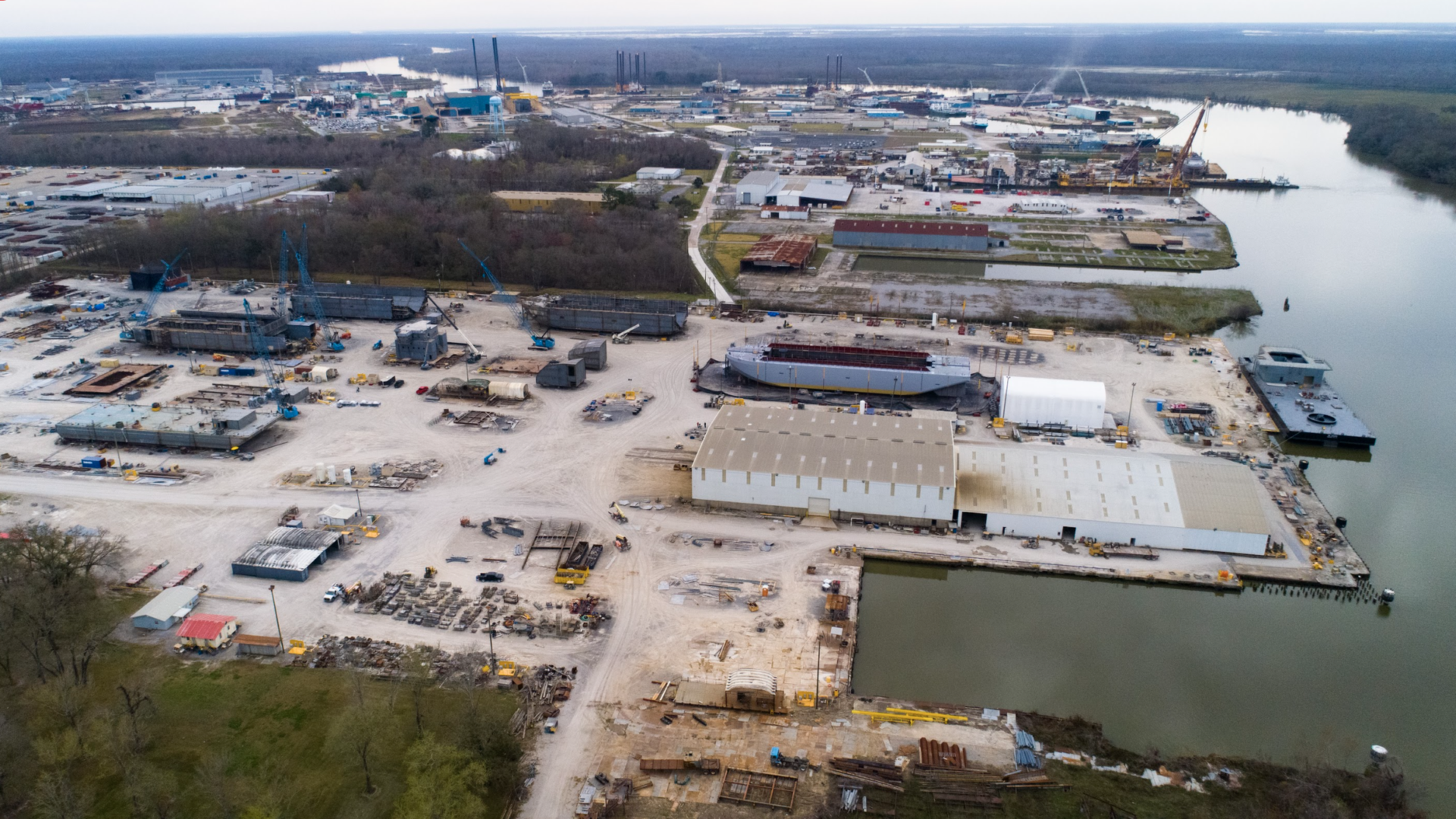 An aerial view of a shipyard. Water is seen on the right. Land and heavy machinery is seen on the left. The sky overhead is gloomy.