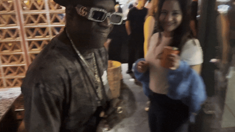 Flavor Flav raises a rhinestone encrusted clock and gestures to it with one hand as a woman behind him prepares to toast a drink but walks away instead.