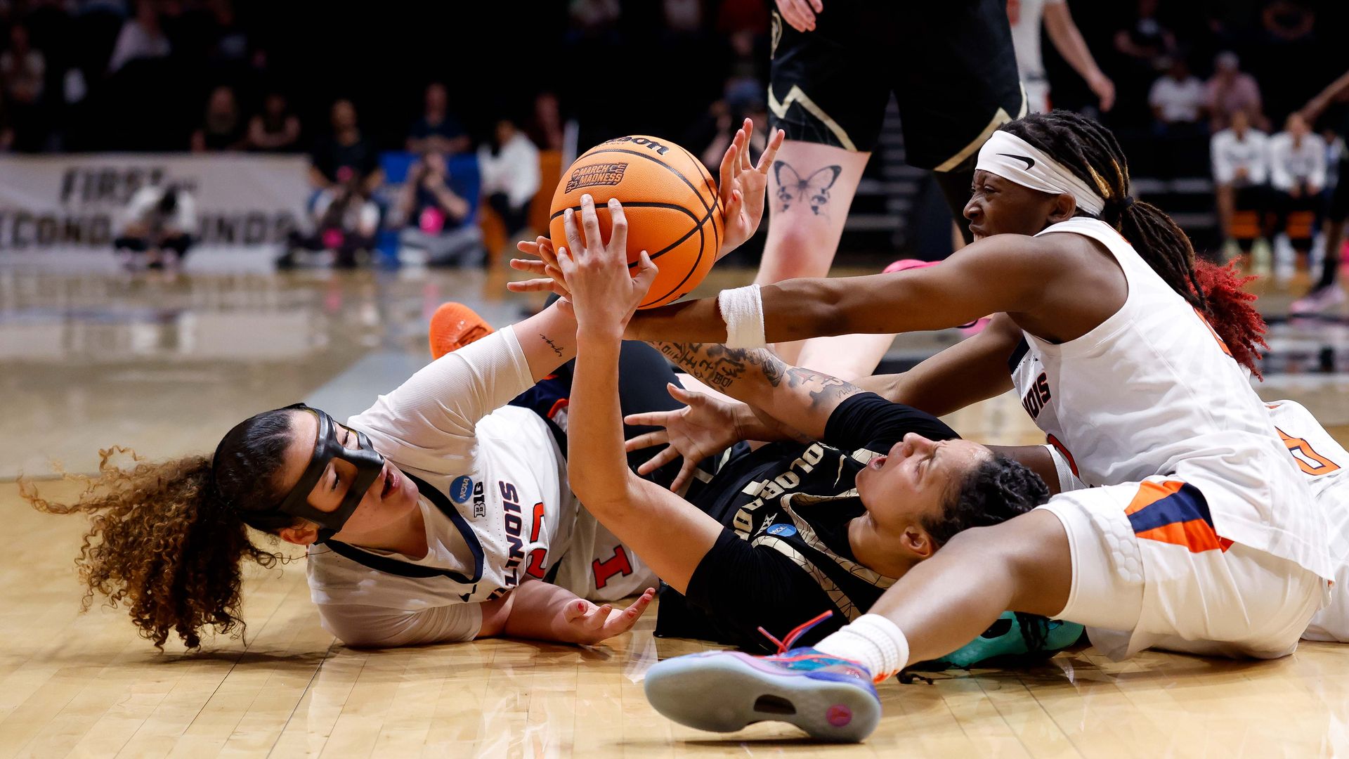 Three female basketball players scramble on the polished court for a loose orange ball during a game; two wear white uniforms with orange accents and one in black, with a crowd in the stands.