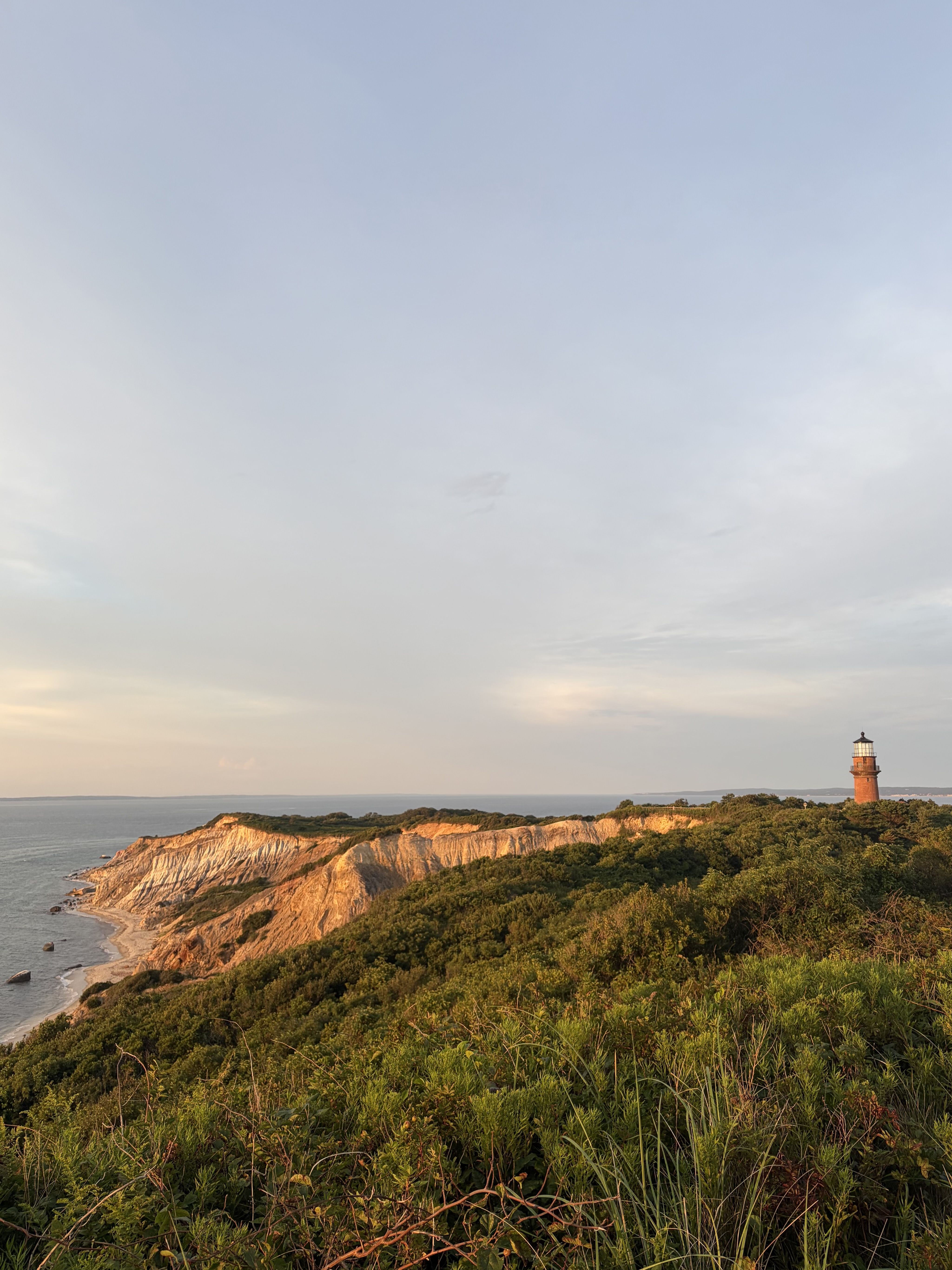 Cliffside coastline with green vegetation and rocky cliffs along the calm sea; a red lighthouse sits on the right, under a pale blue sky.