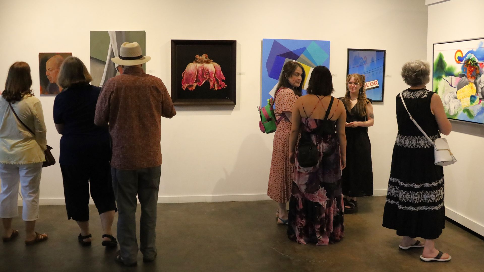 People in an art gallery view various paintings, including portraits, a pink flower, geometric shapes, and a colorful abstract artwork on white walls with a concrete floor.