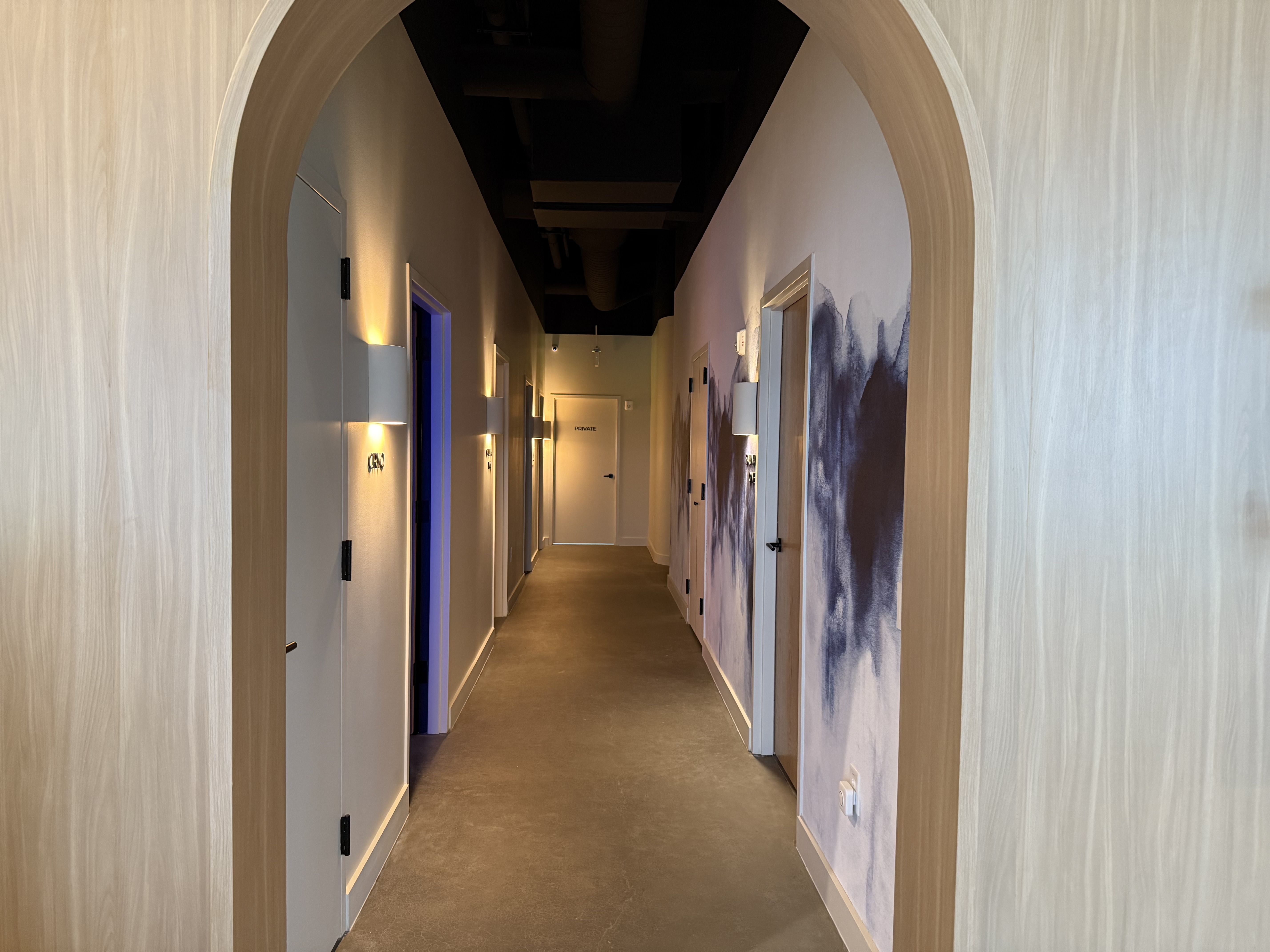 Long hallway with wooden arch entry, warm wall sconces, white doors, a private room door at end, and dark abstract wall art on right side under soft lighting.