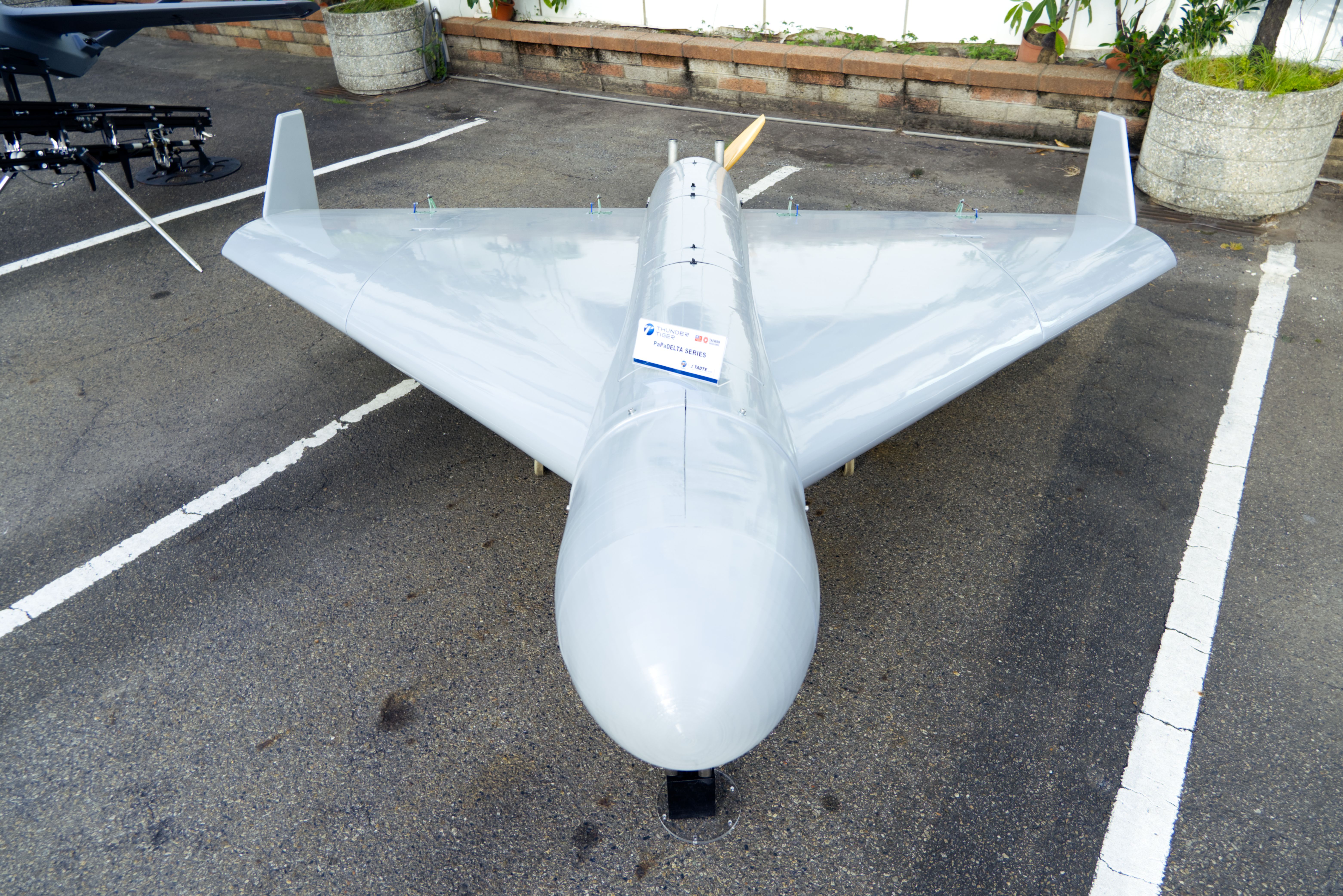 Top-down view of a large silver model airplane parked on an asphalt lot. It has a long fuselage, wide wings, twin vertical tails, and a yellow rear propeller; a label hangs from the fuselage.