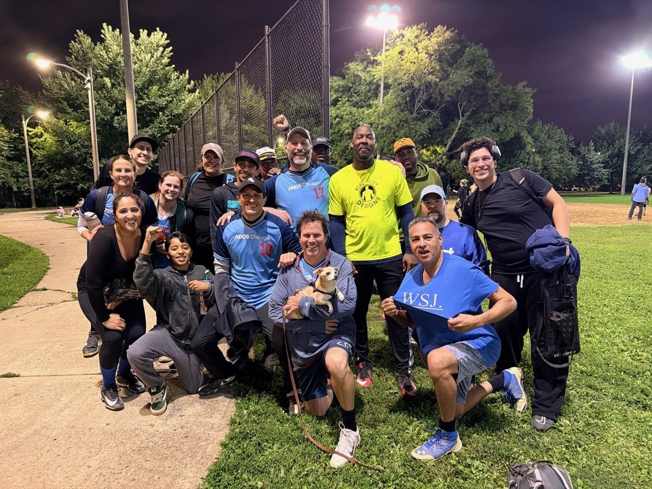 Nighttime group photo of a diverse, smiling team posing near a chain-link fence at a lit baseball field; many wear blue shirts, some in yellow/green, one man holds a small dog.