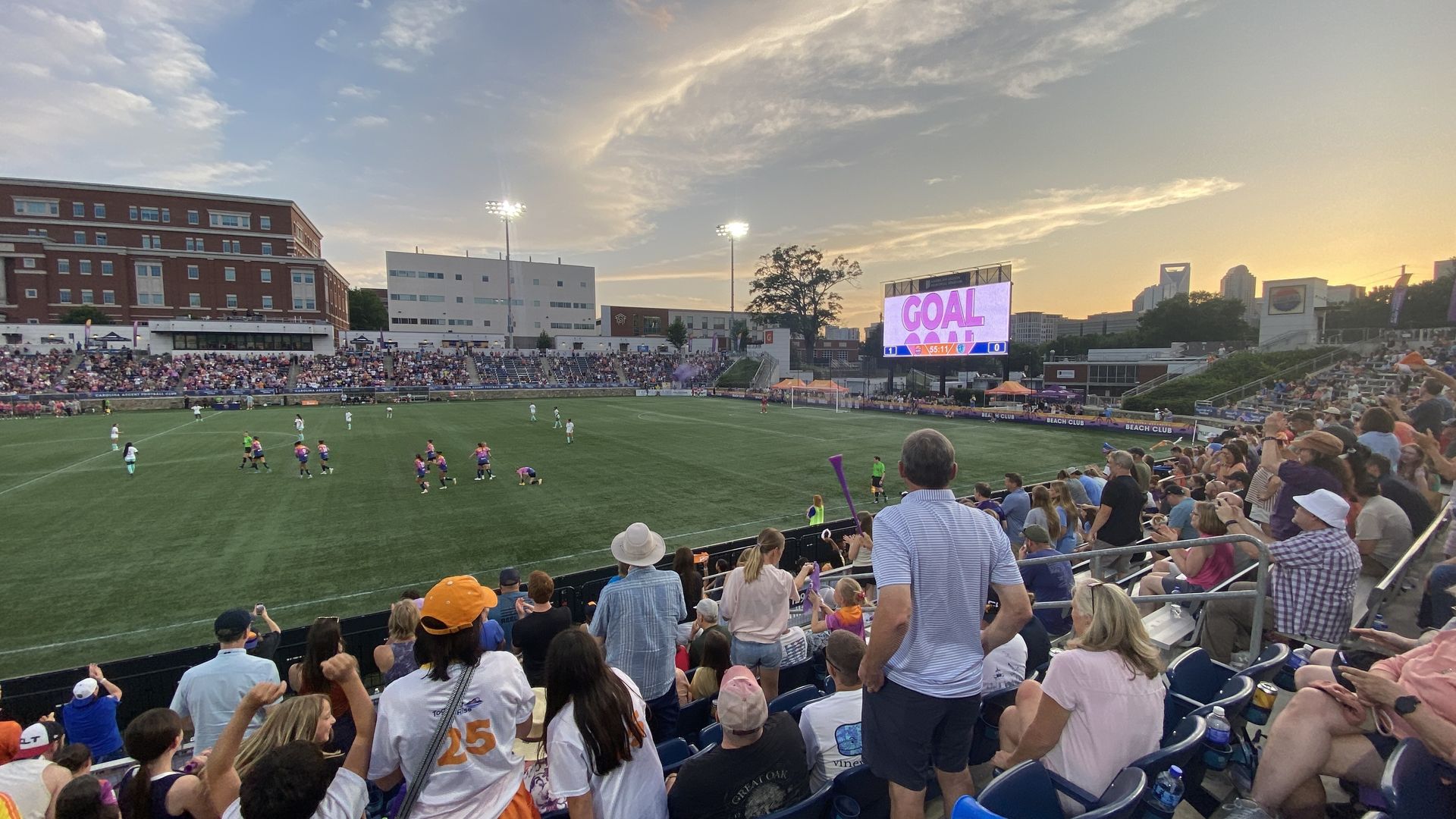 Sunset over a soccer stadium filled with fans; players on the green pitch near the goal, a large scoreboard reading "GOAL", and a city skyline in the background.