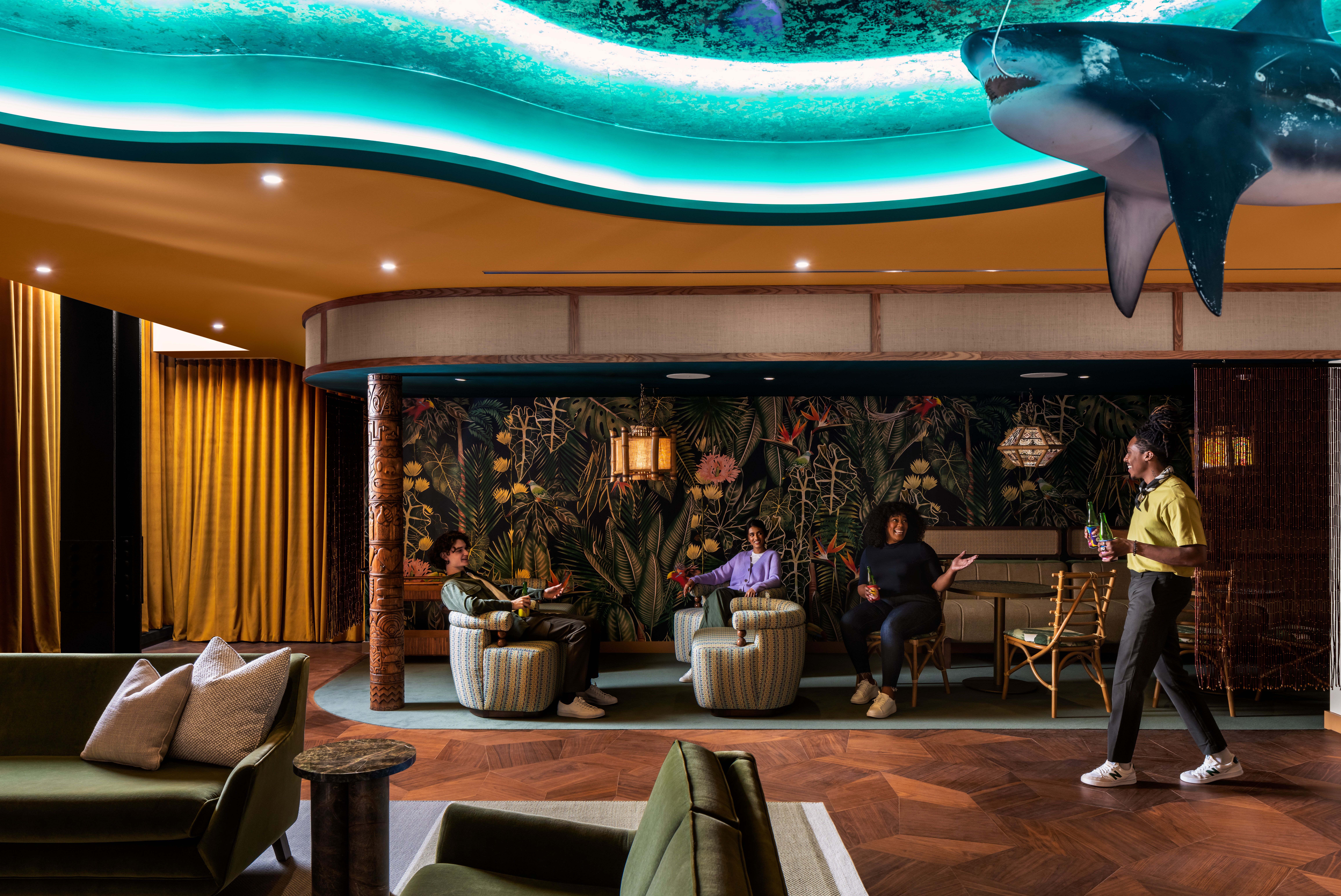 Employees greet a coworker in a tiki lounge with a shark suspended from the ceiling